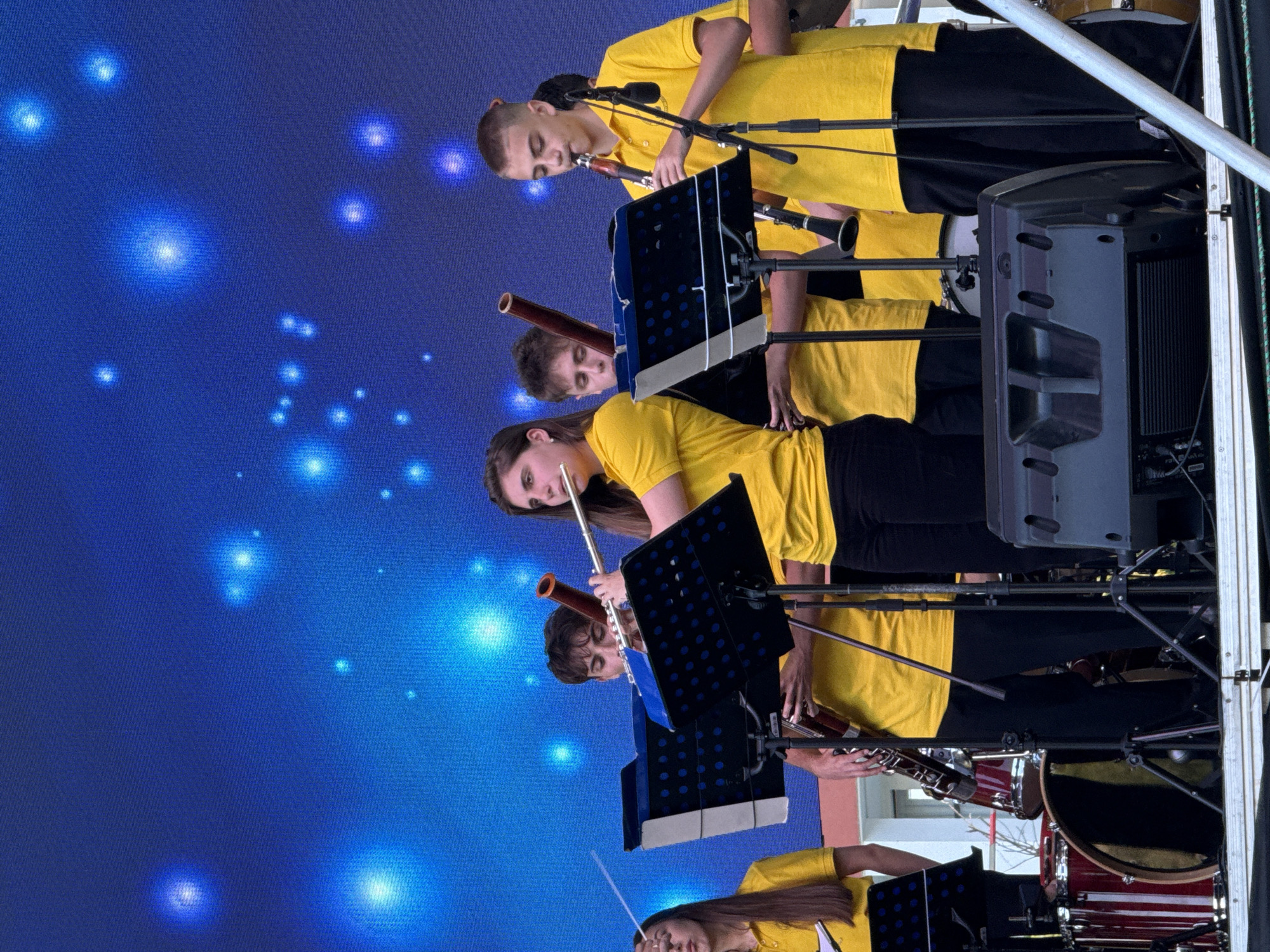 The image shows a youth orchestra performing on a stage. Several young musicians, dressed in yellow polo shirts and black pants, are playing various instruments including flutes, clarinets, and what appears to be a bassoon. They are standing behind music stands displaying sheet music. The background is a large blue screen with bright, bokeh-like lights, simulating a starry or glowing effect. In the foreground, there are audio speakers and parts of a drum set, suggesting an outdoor or large venue performance. The overall scene depicts a moment of musical performance by a young ensemble.