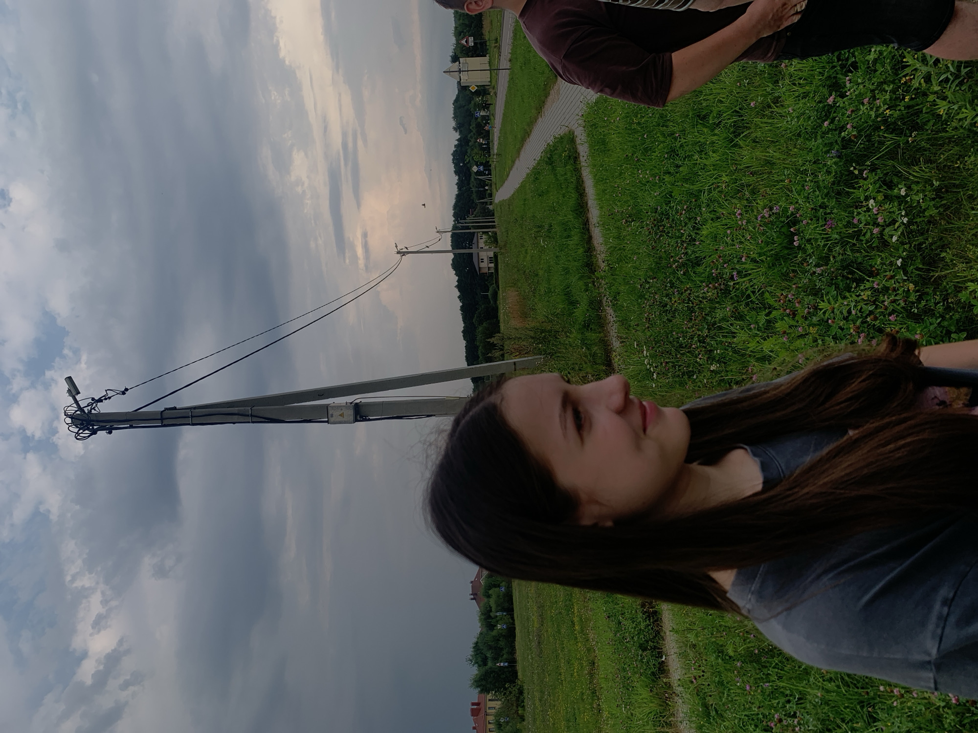 In the foreground, a young female with long, dark hair is captured from the shoulders up, looking to the right with a slight smile. She wears a dark grey t-shirt. To her right, partially visible, is a person wearing a dark maroon t-shirt and black shorts. They are standing on a patch of grass dotted with small purple and white wildflowers.
The background features a paved pathway winding through a grassy area with scattered trees and buildings. A concrete utility pole with electrical wires stretches diagonally across the sky. The sky itself is overcast with heavy, dark clouds, suggesting impending rain or a stormy atmosphere, with some lighter patches indicating dusk or dawn. The overall environment suggests a suburban or rural outskirts area. The presence of buildings and a paved road indicates it is a populated area.