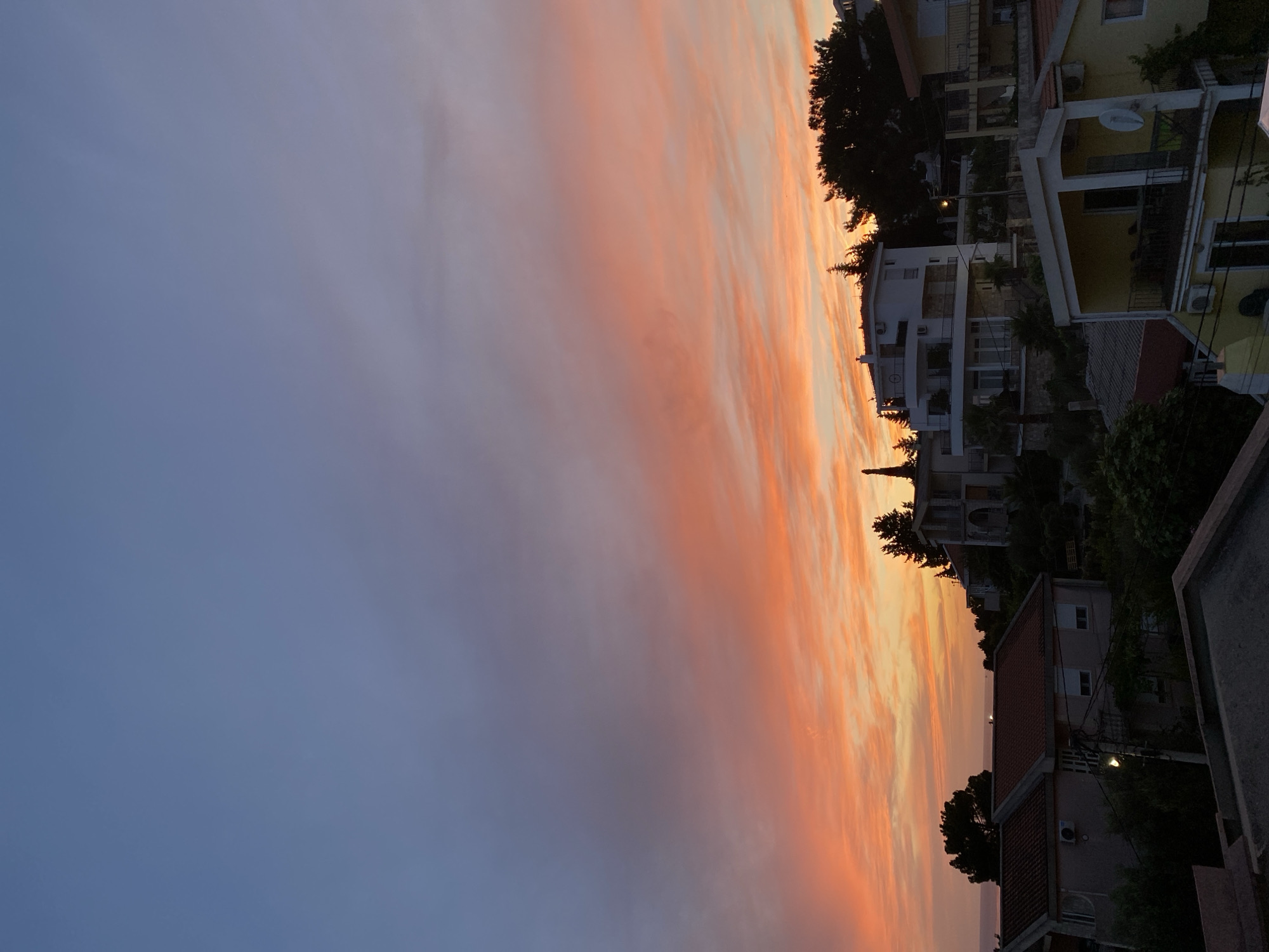 This image depicts a sunset over the town of Šušanj, Montenegro. The sky is a gradient of colors, starting with a pale blue at the top and transitioning to vibrant orange and pink hues near the horizon. Wispy clouds are visible, catching the light of the setting sun.
In the foreground and mid-ground, several buildings are visible. These appear to be houses or villas, some with red-tiled roofs and others with lighter colored exteriors. They are situated on a hillside, with lush green vegetation surrounding them. The architecture suggests a Mediterranean style, with balconies and terraces visible on some structures.
In the far background, a body of water, likely the Adriatic Sea, can be seen. The distant coastline is dimly illuminated by the sunset.
The image is a still photograph, capturing a serene moment at dusk. There are no people visible in the frame. The overall impression is one of natural beauty and peacefulness.