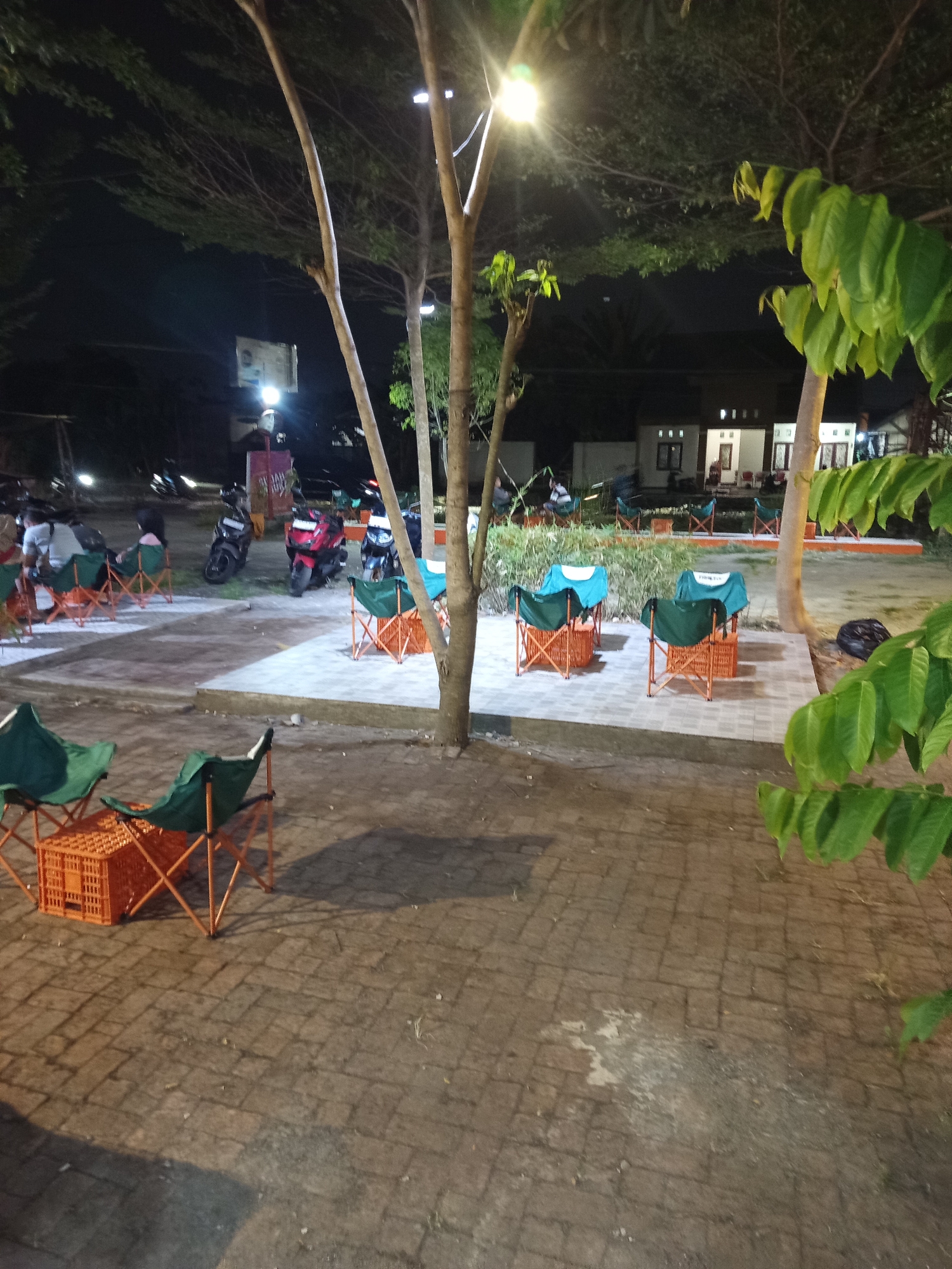 At night in the city of Becirongengor, Indonesia, an outdoor seating area is depicted. The scene is illuminated by artificial lights, casting shadows on the brick-paved ground. Several portable chairs, primarily green with orange frames, are arranged around orange plastic crates that serve as makeshift tables. Some of these chairs are occupied by individuals who are partially visible. In the background, a row of motorcycles is parked, suggesting a public gathering or establishment. Beyond the seating area, a building with windows and a roof is visible, indicating the presence of structures in the vicinity. Trees with dense foliage are scattered throughout the scene, some of which have lights hanging from their branches. The overall atmosphere is casual and appears to be a relaxed outdoor setting at dusk or after dark.
