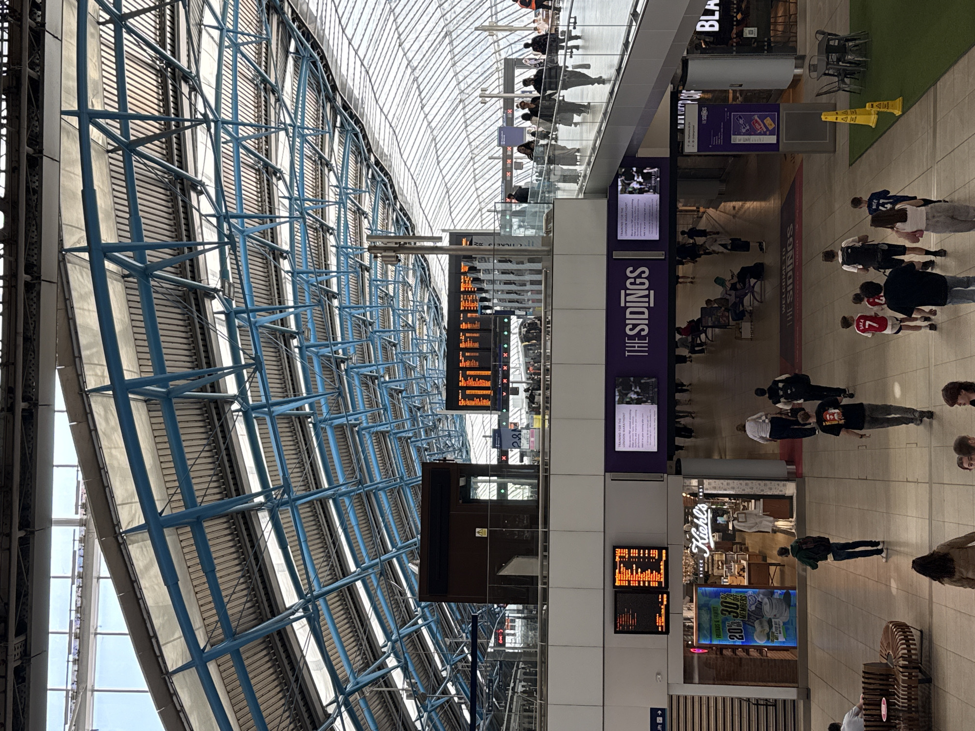 The image depicts the interior of a transportation hub, potentially a railway station, in London, United Kingdom. The architectural structure includes a vast, high-ceilinged space with a complex network of blue steel beams supporting a glass roof. Passengers are walking along the concourse, some are seated and waiting. There are shops such as "Kiehl's" and "The Sidings". Electronic display boards show train departure information. People are wearing casual clothing. The image captures a typical scene of transit and commerce in a modern transportation hub.