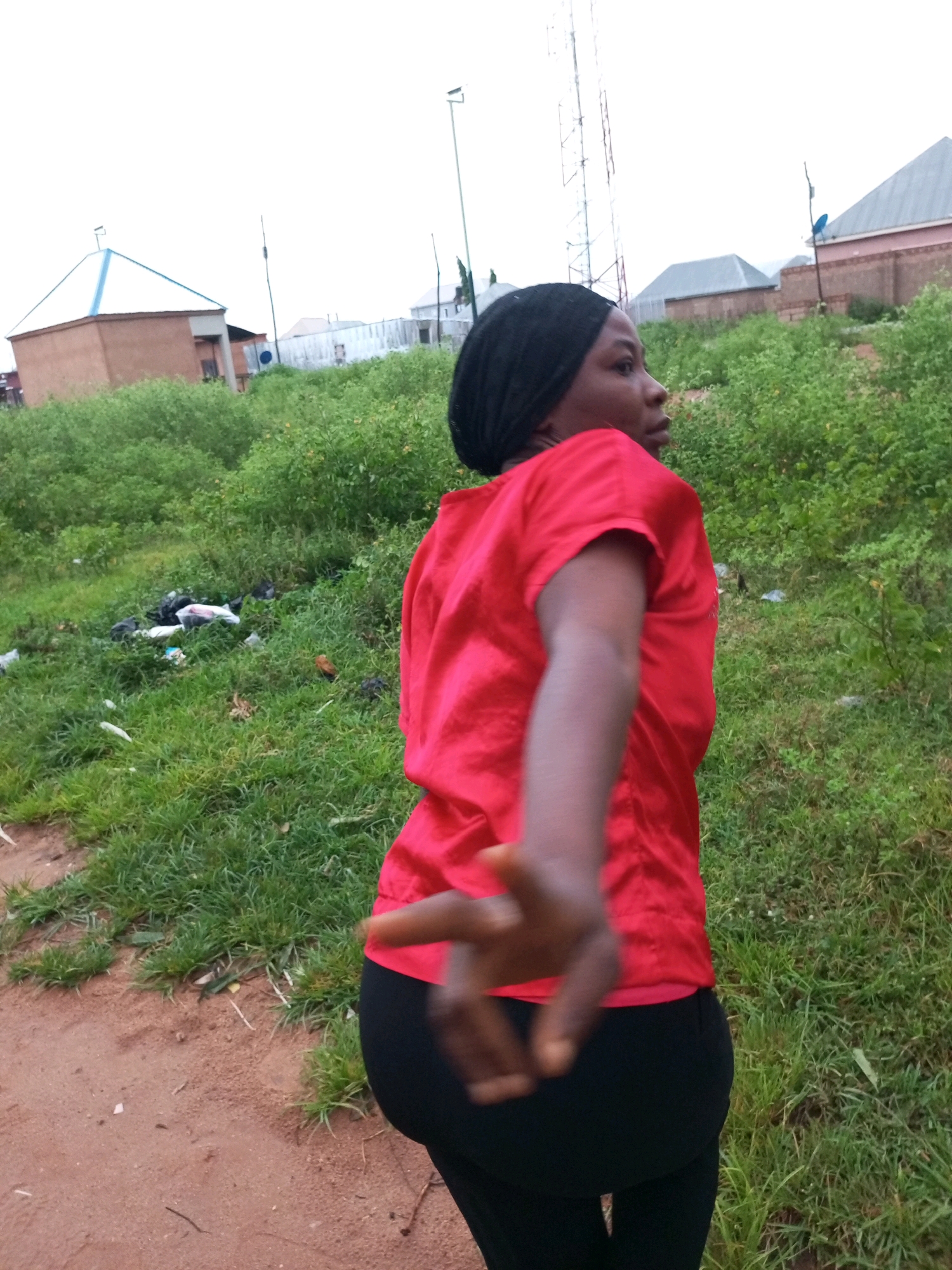 A young woman wearing a black headscarf and a red shirt is captured in a candid, outdoor shot in Tempure, Nigeria. She is positioned facing away from the camera, with her right arm extended towards the viewer in a gesture that suggests she is either reaching out or posing. Her left side is partially visible, and she appears to be wearing black pants.
The environment is somewhat unkempt, with a patch of green grass and some scattered debris visible in the background. Beyond the vegetation, several buildings and a tall telecommunications tower can be seen, hinting at an urban or semi-urban setting within Tempure. The sky above is overcast, contributing to a diffused lighting condition. The image appears to have been taken from a low angle, emphasizing the woman and her surroundings.