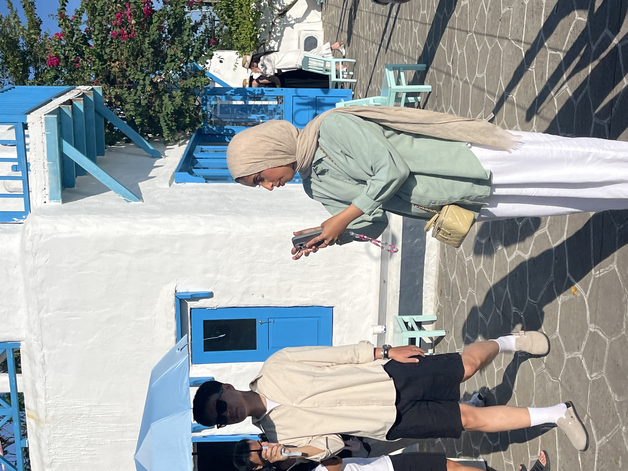 The image captures a scene in Đà Nẵng, Vietnam, characterized by a white building with blue trim. A woman wearing a headscarf and a light green shirt is looking at her phone. A man wearing black shorts, a light tan shirt, and sunglasses is also present. The ground is paved with stone. There are chairs and other people in the background. Bougainvillea is visible in the upper left corner. The scene is brightly lit, suggesting daytime.