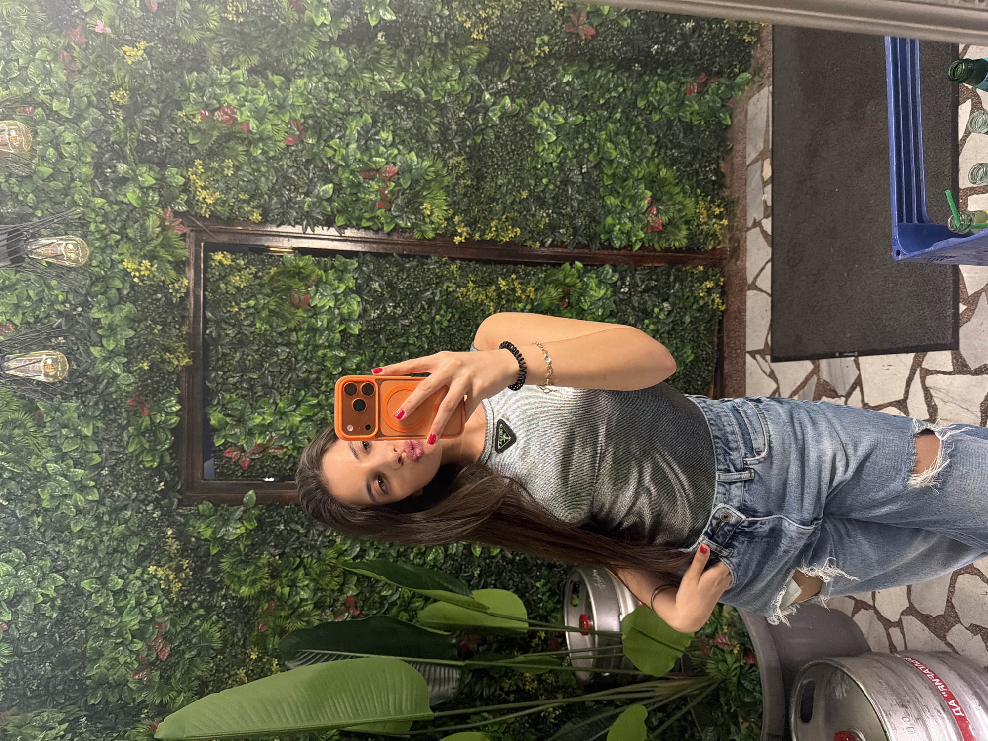 A young woman is taking a selfie in front of a green wall adorned with lights and framed by dark wood. She is wearing a gray ribbed tank top and ripped blue jeans. She is holding an orange smartphone, likely to capture the image. The background includes green plants, possible beer kegs, and a patterned floor. There is a reflection visible in the background, showing a blue object. The location is Rudartsi, Bulgaria.