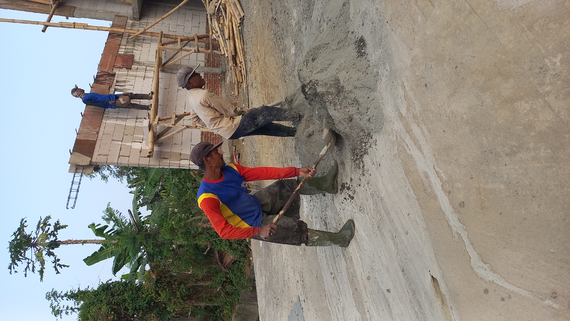 In Mangunsari, Indonesia, a construction site is depicted with three male workers actively engaged in their tasks. In the foreground, two men are shoveling a pile of concrete mix. One worker, wearing a red, yellow, and blue long-sleeved shirt, dark shorts, and green boots, is bent over, using a shovel to move the material. To his right, another man, dressed in a light-colored long-sleeved shirt and dark pants, is also shoveling the same concrete mix. In the background, a third worker, wearing a blue long-sleeved shirt and dark pants, stands on a wooden scaffolding near a partially constructed brick wall. The environment is characterized by construction materials, including bamboo poles and rebar, and lush green foliage surrounds the site, with a tall tree in the background. The sky above is clear and pale.