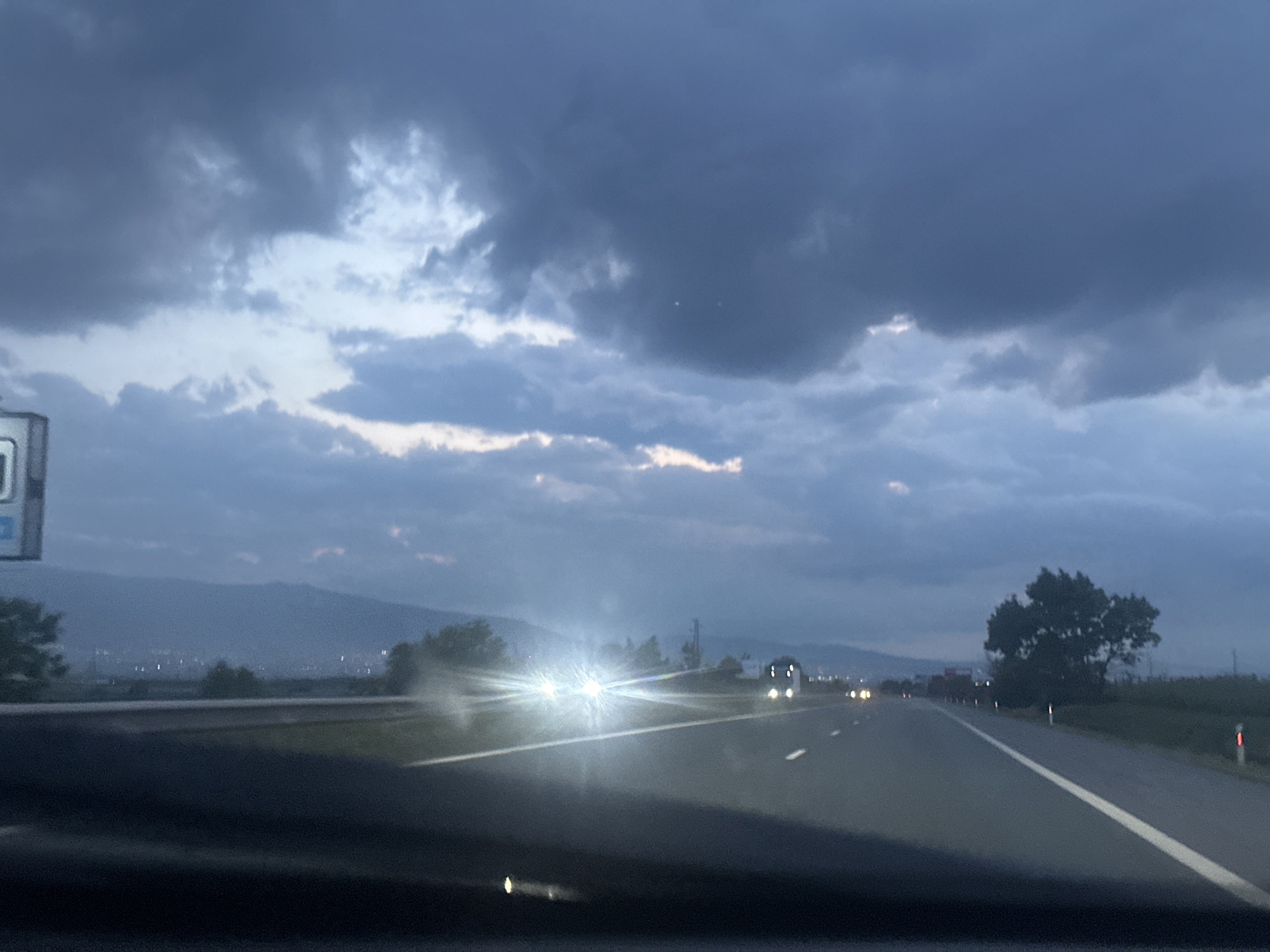 This media file captures a view from inside a vehicle, looking out onto a highway near Zhelyava, Bulgaria, at dusk or twilight. The upper half of the image is dominated by a dramatic, overcast sky filled with dark, heavy clouds, with lighter segments visible between them, suggesting low light and potentially stormy weather. The highway extends into the distance, featuring white lane markings. Bright, blurred headlights from oncoming vehicles are prominent in the middle ground, indicating active traffic and low visibility. Other vehicles appear as faint shapes or illuminated taillights further along the road. In the background, hazy mountains or hills are visible, with scattered lights at their base on the left, hinting at a distant town or city. To the right of the road, a dark, prominent tree stands, alongside other vegetation and red and white reflective road markers. The foreground shows a partial view of the vehicle's dashboard. No legible text is visible on any signs or objects within the frame. The image conveys a sense of evening travel under a dramatic and moody sky.