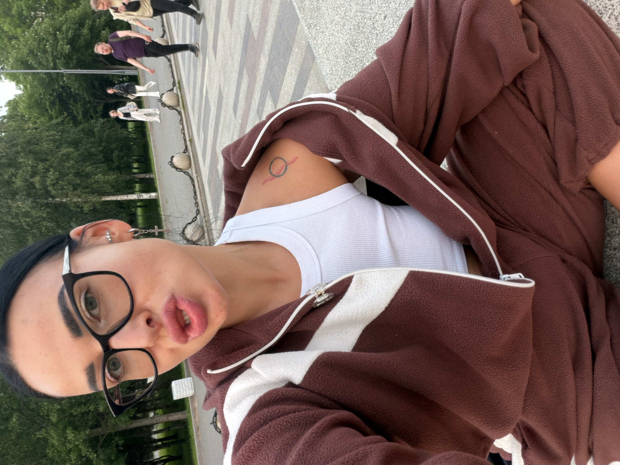 This media file is a close-up selfie, rotated 90 degrees clockwise, featuring a young person resting outdoors in what appears to be a park or public pathway in Saint Petersburg, Russia.
The subject, seen from the chest up, has dark hair, dark eyebrows, and wears black-rimmed glasses that frame green eyes. Their expression is characterized by a "duck face" or pout. They have multiple ear piercings, including a small cross earring dangling from the right ear. They are wearing a white ribbed tank top layered under a brown fleece jacket with white stripes and a visible white zipper. On their upper left arm, a small, minimalist tattoo is visible, depicting a black circle with a red wavy line underneath.
In the background, visible over the subject's shoulder and to their left (in the original, rotated orientation), several people are walking on a paved pathway with a geometric pattern. A low barrier consisting of stone posts connected by metal chains separates the pathway from a grassy area with numerous green trees. The overall setting suggests a pleasant daytime scene, likely overcast or during the early morning/late afternoon, given the soft natural light. No specific activities beyond casual walking are discernible among the background figures. No visible text is present in the image.