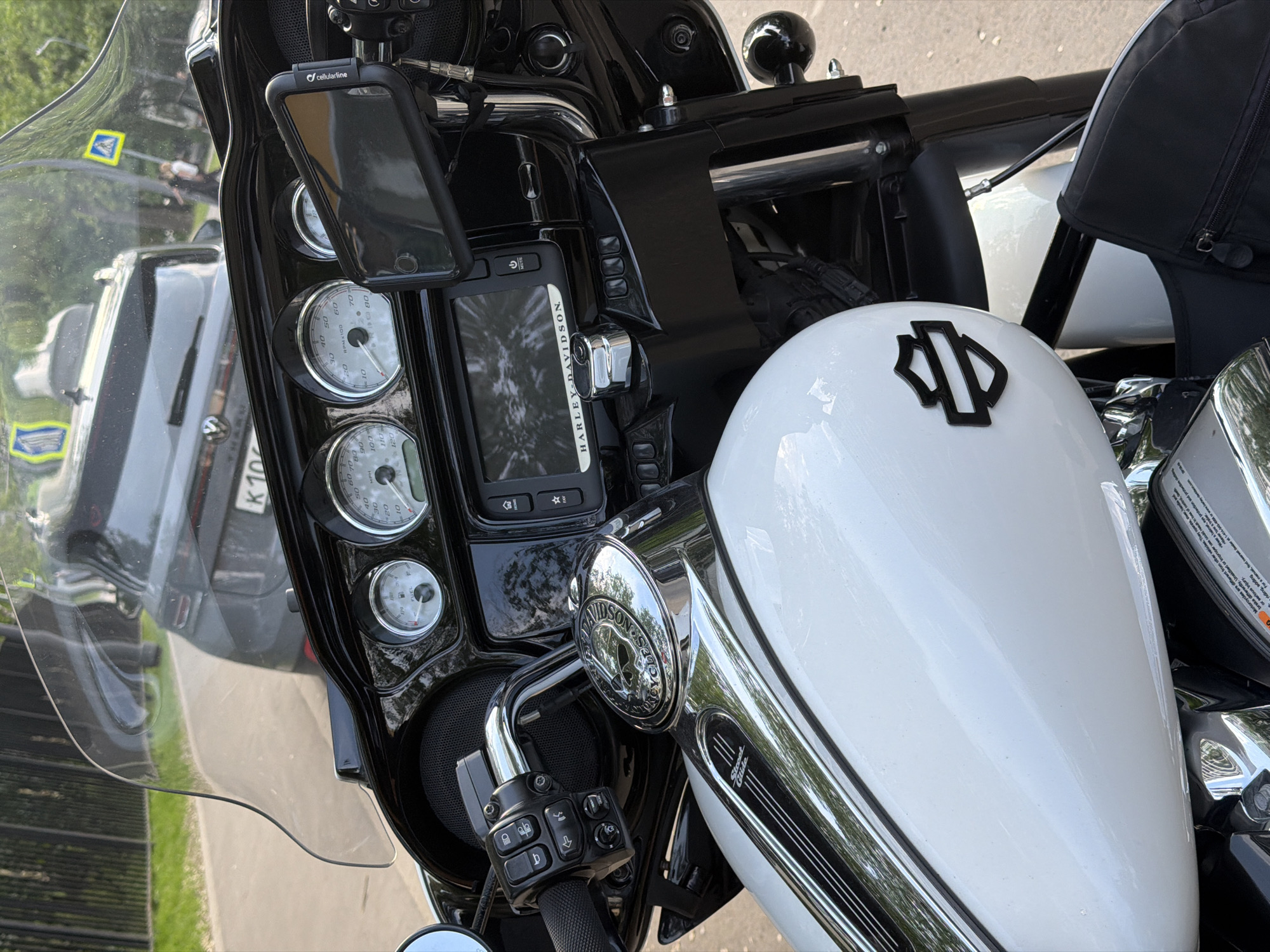 The image shows a close-up of the dashboard and fuel tank of a Harley-Davidson Street Glide motorcycle. The motorcycle has a white fuel tank with the Harley-Davidson logo. The dashboard has a speedometer, tachometer, and other gauges, along with a phone mount and a GPS device. The image also includes a partial reflection of a car with a "K100" license plate, suggesting it was taken in Moscow, Russia. The motorcycle is likely parked on a street or sidewalk.