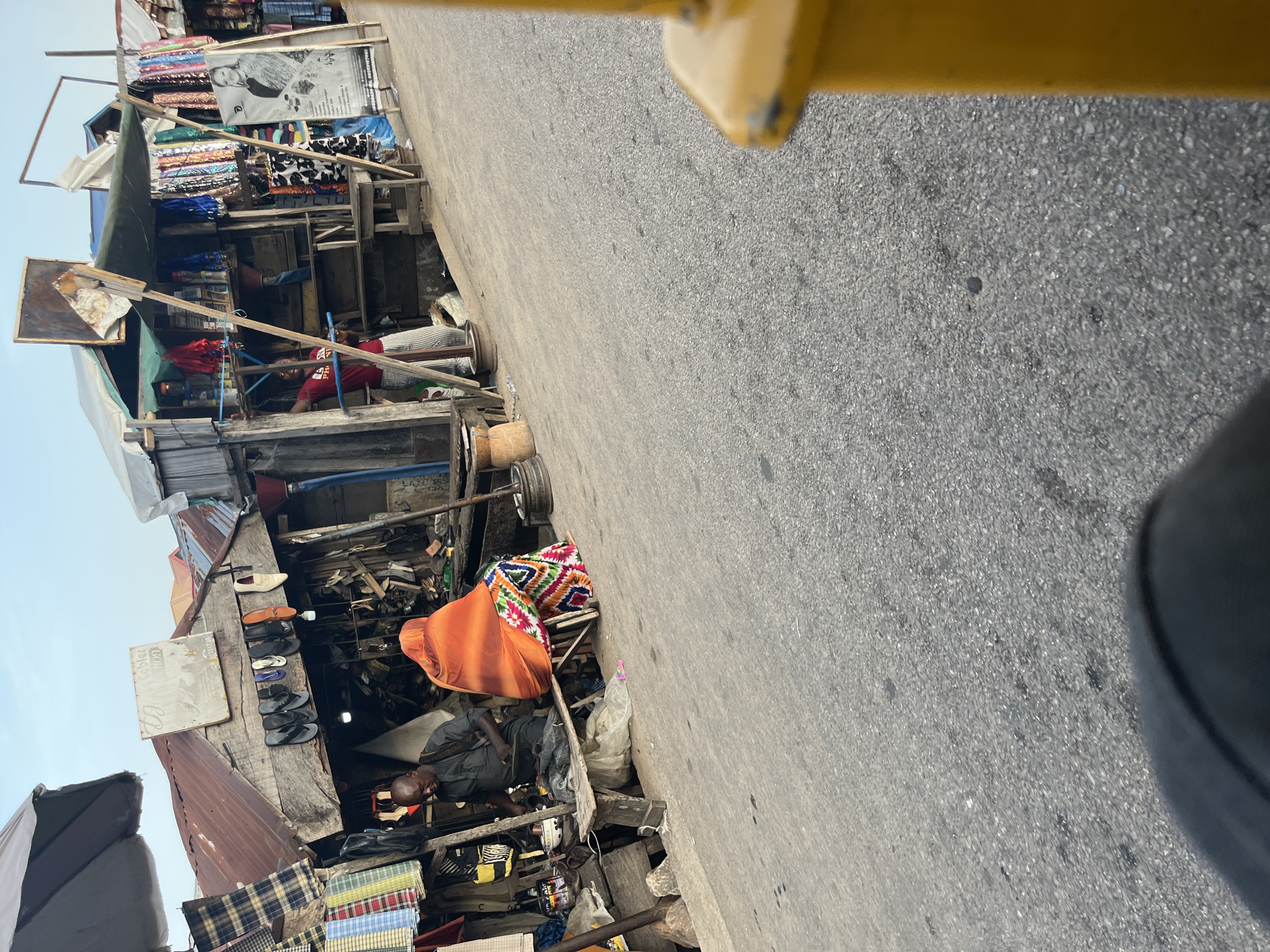 An outdoor market scene shows a man sitting on a wooden bench, working on shoes. He is wearing a dark shirt and has a bag slung over his shoulder. Beside him, a person in an orange headscarf and a brightly colored patterned skirt sits on a stool. Behind them, various stalls are set up with goods displayed, including shoes hanging from a wooden structure, fabrics, and other merchandise. A person in a red shirt and checkered pants stands in a stall to the right, appearing to be interacting with someone off-camera. The stalls have corrugated metal roofs and wooden framing. The ground is paved asphalt. The sky is visible in the background, with a slightly hazy, light blue hue. A yellow object is partially visible in the foreground on the right.