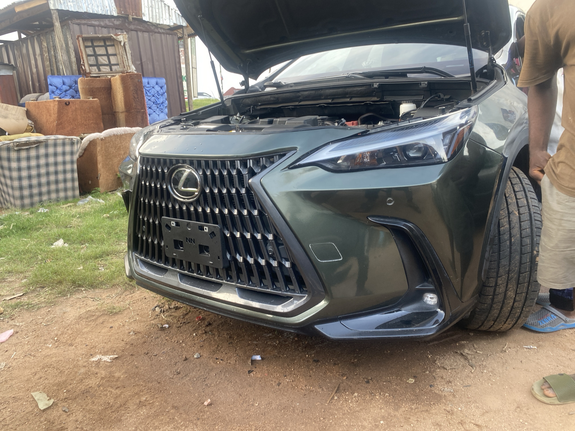 A dark green Lexus SUV is parked on a dirt road in Jalingo, Nigeria. The hood of the SUV is open, revealing the engine. A person wearing a brown t-shirt and shorts is standing next to the driver's side of the vehicle, with their right arm resting on the fender. Another person, only their legs and feet are visible, is standing behind the SUV wearing blue slides and green flip-flops. In the background, there are several stacks of discarded furniture and building materials, including a corrugated metal structure, wooden crates, and blue upholstered cushions. The ground is covered in dirt and patches of dry grass.