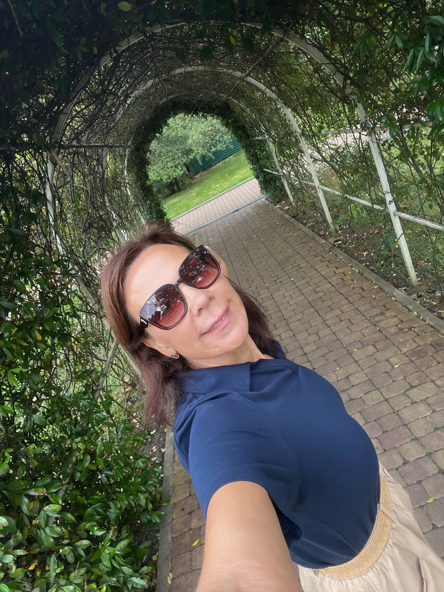 A woman with dark brown hair and sunglasses is taking a selfie in a park. She is wearing a navy blue collared shirt and a light-colored skirt. The background shows a pathway made of brick pavers, a green hedge, and a trellised archway covered in vines. The overall setting appears to be an outdoor garden or park area, possibly in Sochi, Russia, given the mention of the city. The lighting suggests it is daytime.