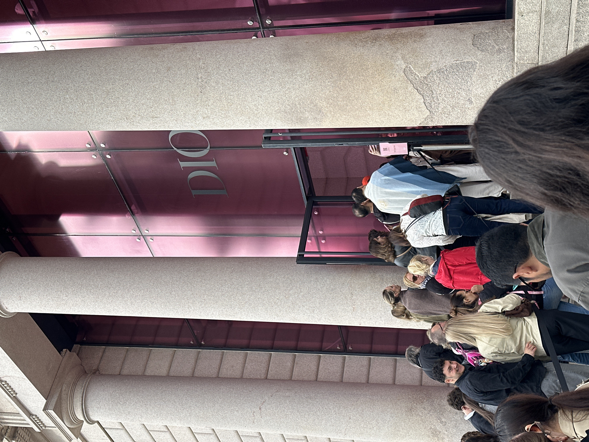 A street-level view of the front of a DIOR store in Milan, Italy. A line of people are waiting to enter the store. The storefront is made of glass and metal. The word "DIOR" is written vertically on the store facade. The facade also features beige stone columns and steps. People in line are looking at the entrance. Most of the people in line are looking at the entrance and some are conversing.