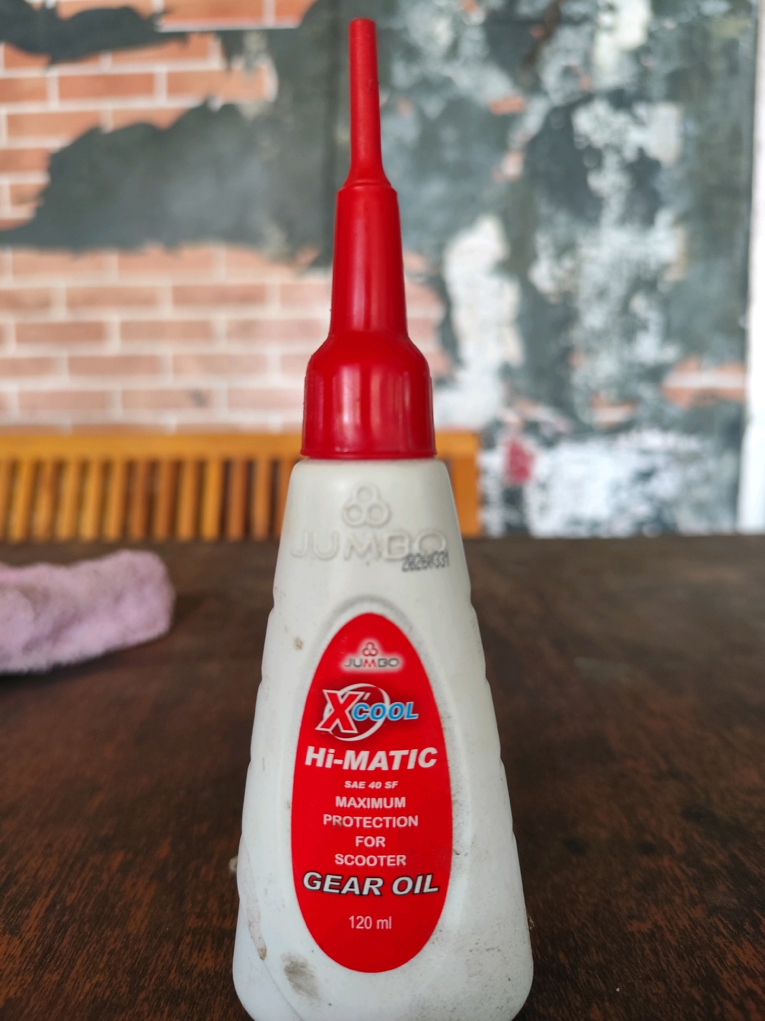 A white plastic bottle of "X-COOL Hi-MATIC GEAR OIL" with a red applicator tip stands on a wooden surface. The bottle has a red label with white text. The background shows a brick wall with some patchy plaster, and a blurred view of what appears to be a wooden bench. The scene suggests an indoor setting with natural light. The image quality is moderate, with some dust and wear visible on the bottle. No people are present.