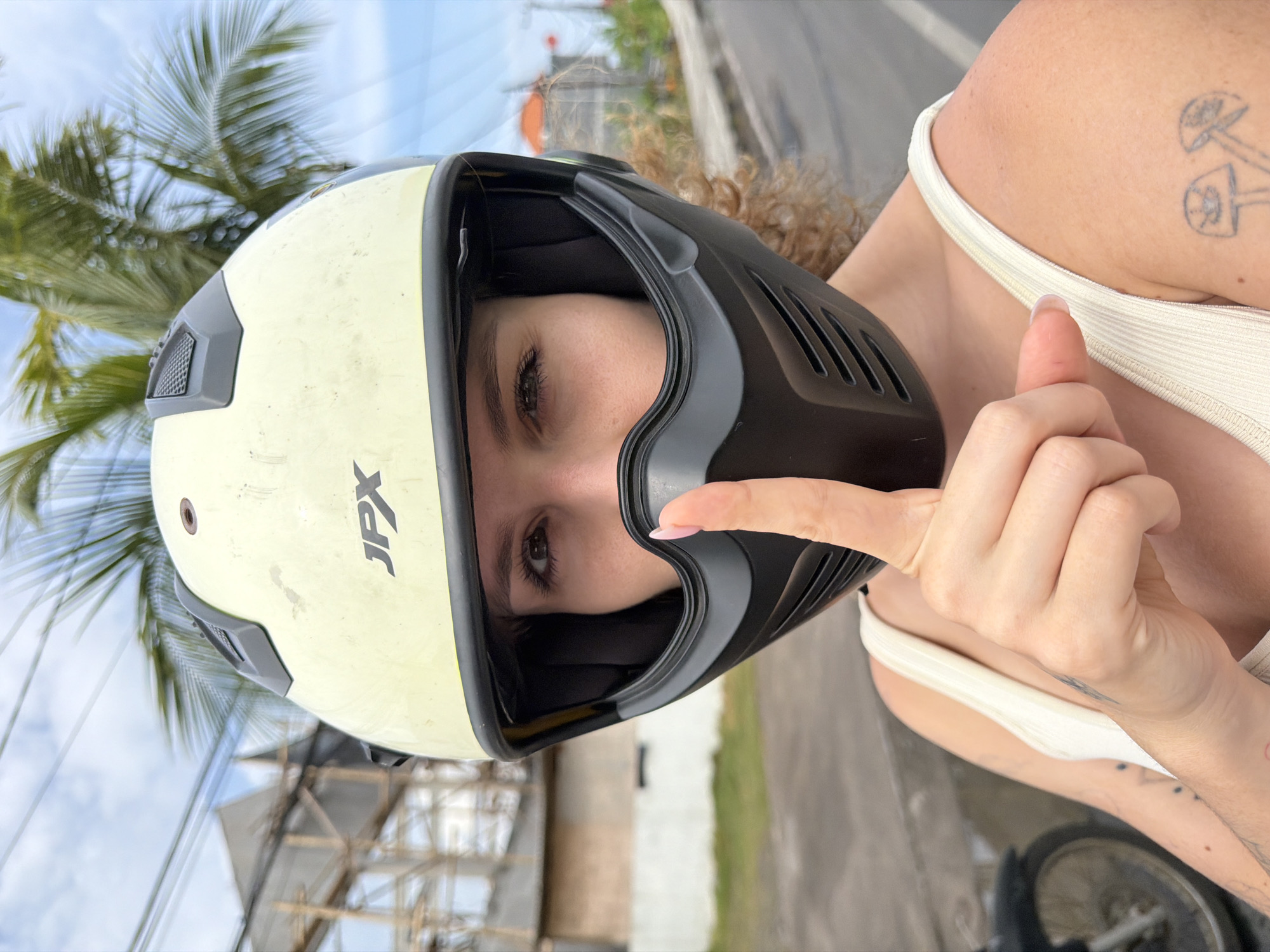 A woman wearing a cream-colored helmet with black accents and a black face mask is holding her index finger to her lips in a "shushing" gesture. The helmet has the "JPX" logo on the front. She is wearing a white tank top. Her left arm is visible, showing tattoos of mushrooms and other symbols. In the background, palm fronds are visible against a blue sky. There is also a structure with scaffolding and wiring, and a road with a motorbike wheel visible on the left. The overall environment suggests a tropical urban setting. The location is identified as Mengwi, Indonesia.