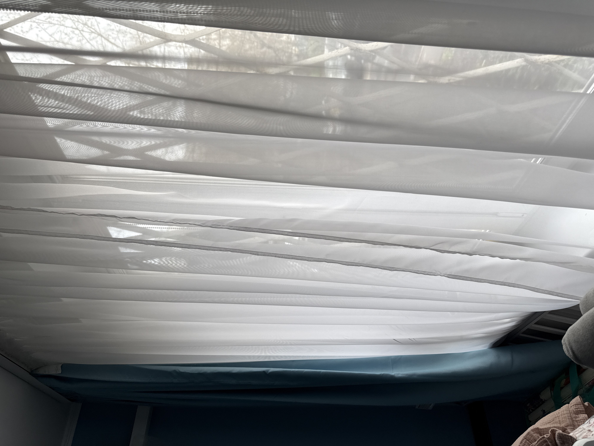 The image depicts the underside of a white fabric window shade or blind. The shade is partially obscuring a window, through which sunlight and the silhouettes of tree branches are visible. Below the shade, a section of a blue-colored mattress or cushion is visible. The context of Moscow, Russia, is not supported by the visual information presented in the image. There are no people, actions, or interactions visible in the frame.