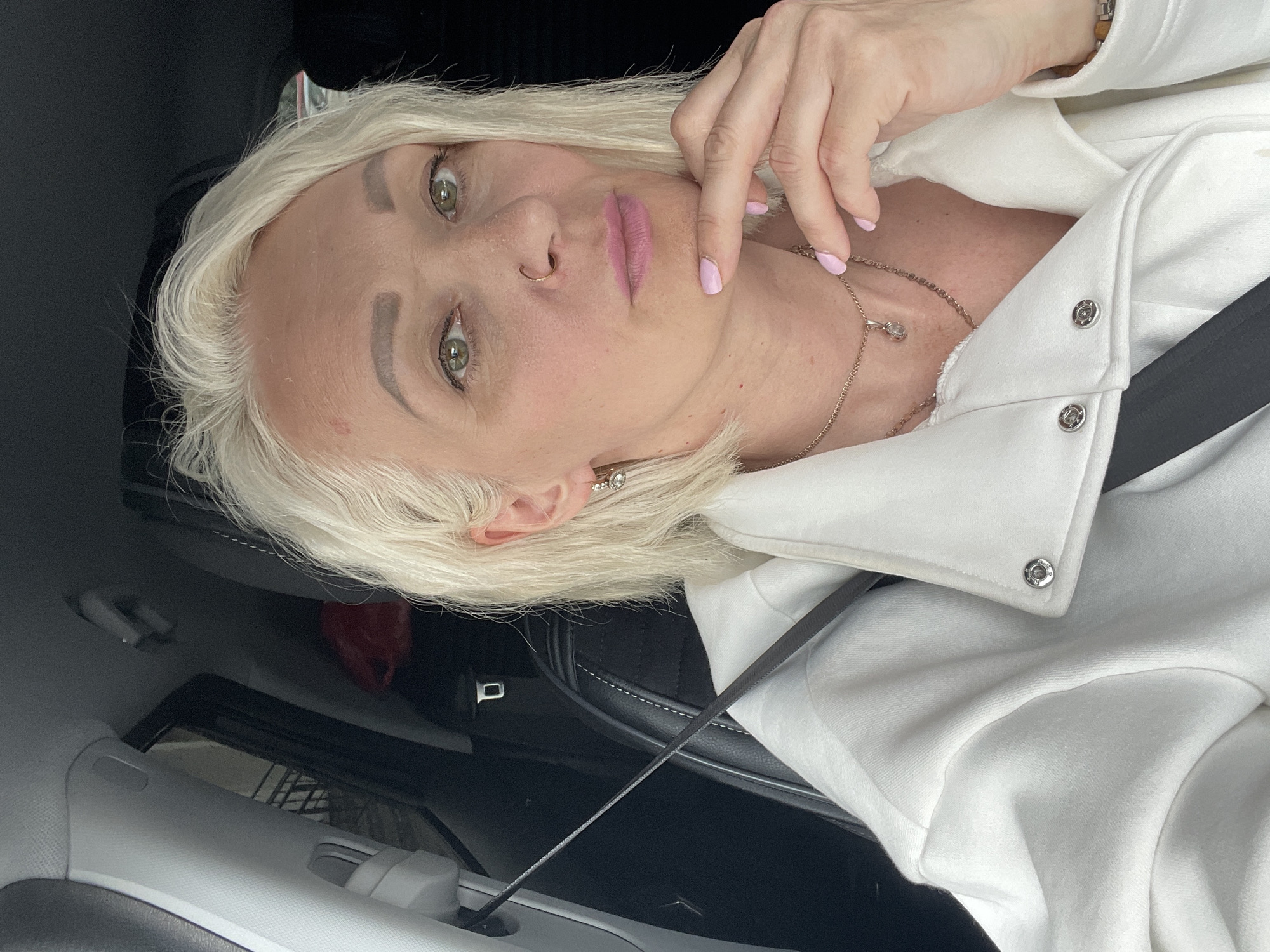 A woman with blonde hair and green eyes is sitting in the driver's seat of a car. She is wearing a white jacket and a necklace. Her hand is resting on her chin. A red bag is visible in the background. The image was taken in the city of Gatchina, Russia.