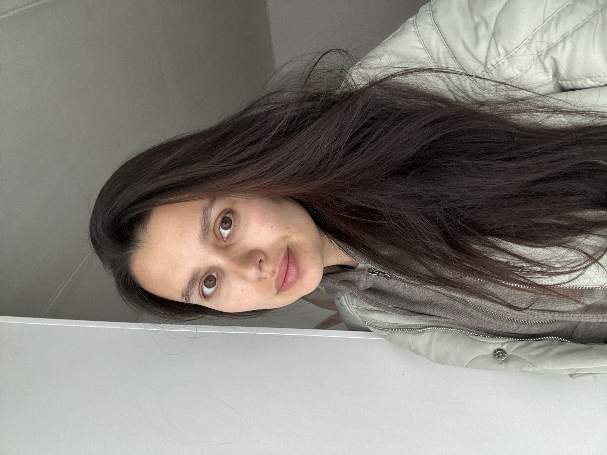 A woman with long, dark hair is facing the camera. She has brown eyes and light skin. She is wearing a gray hooded sweatshirt and a light-colored, quilted jacket. The background is a plain, light gray wall. There is no evidence to suggest the location is Chaikovsky, Russia.