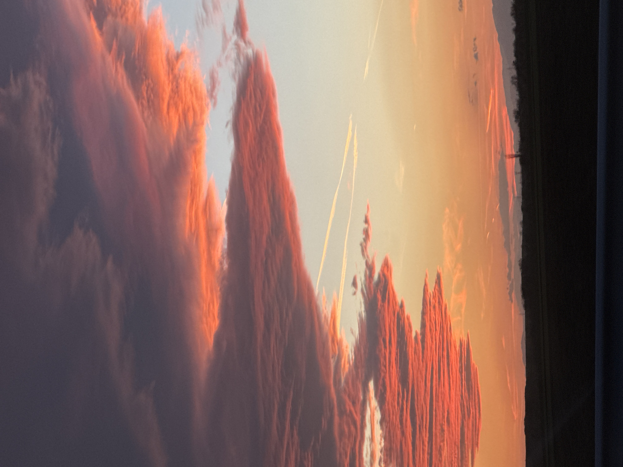 The image shows a sunset sky with orange and red-tinged clouds. Two contrails are visible. The edge of a wooded area is on the horizon. The photograph was taken from indoors, as a curtain is visible. There is no evidence to suggest the photo was taken in Malenovo, Bulgaria.