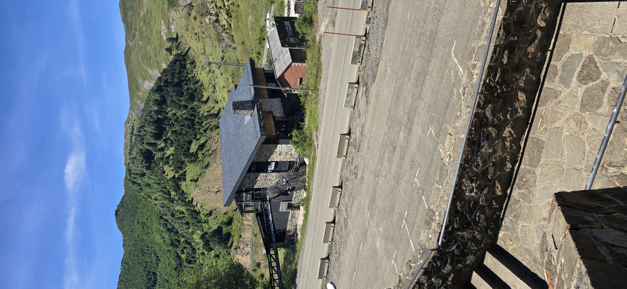The image shows a modern building with a stone facade and a dark, angled roof, located on a mountainside in Sallent de Gállego, Spain. The building is partially obscured by trees and vegetation. A paved road runs in front of the building, with concrete barriers along the edge of the road. There are parking spaces marked on the asphalt. A smaller, red-roofed building is visible next to the main building, with a sign that reads "THE CLUB". The background consists of lush, green, tree-covered mountains under a bright blue sky with a few wispy clouds. In the foreground, a stone-paved staircase with a metal railing leads down out of frame. The scene appears to be a clear, sunny day.