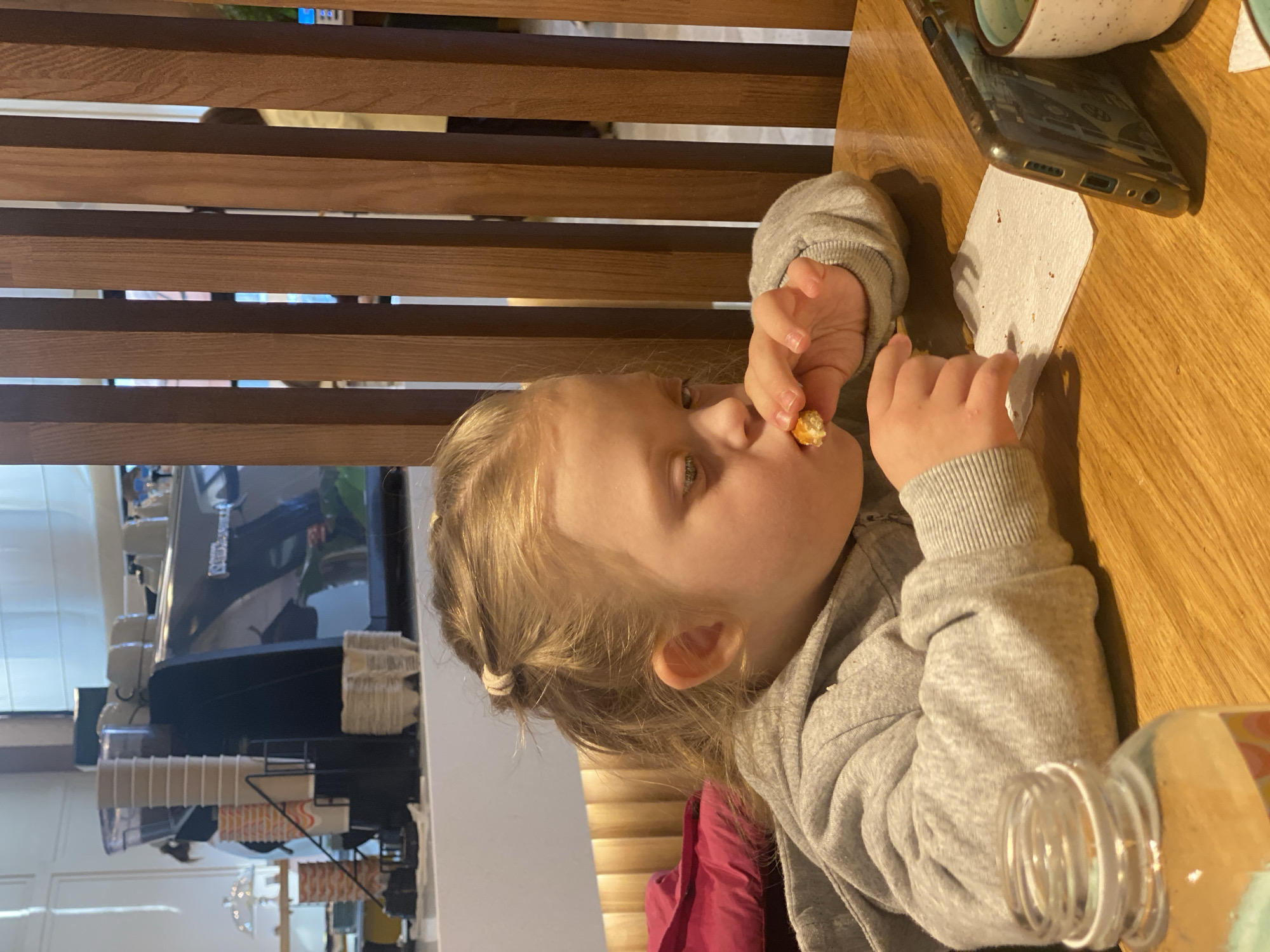 A young child with fair hair, wearing a gray sweatshirt, is seated at a wooden table, eating. The child is holding a small, round, golden-colored piece of food near their mouth. A white napkin lies near their hands. A smartphone is on the table next to the napkin. A glass jar with a lid is positioned near the child. The background shows wooden slats, possibly part of a partition or furniture, and a section of a cafe or restaurant setting with coffee equipment and disposable cups. Given the prompt, this image was most likely taken in a cafe in Yekaterinburg, Russia.