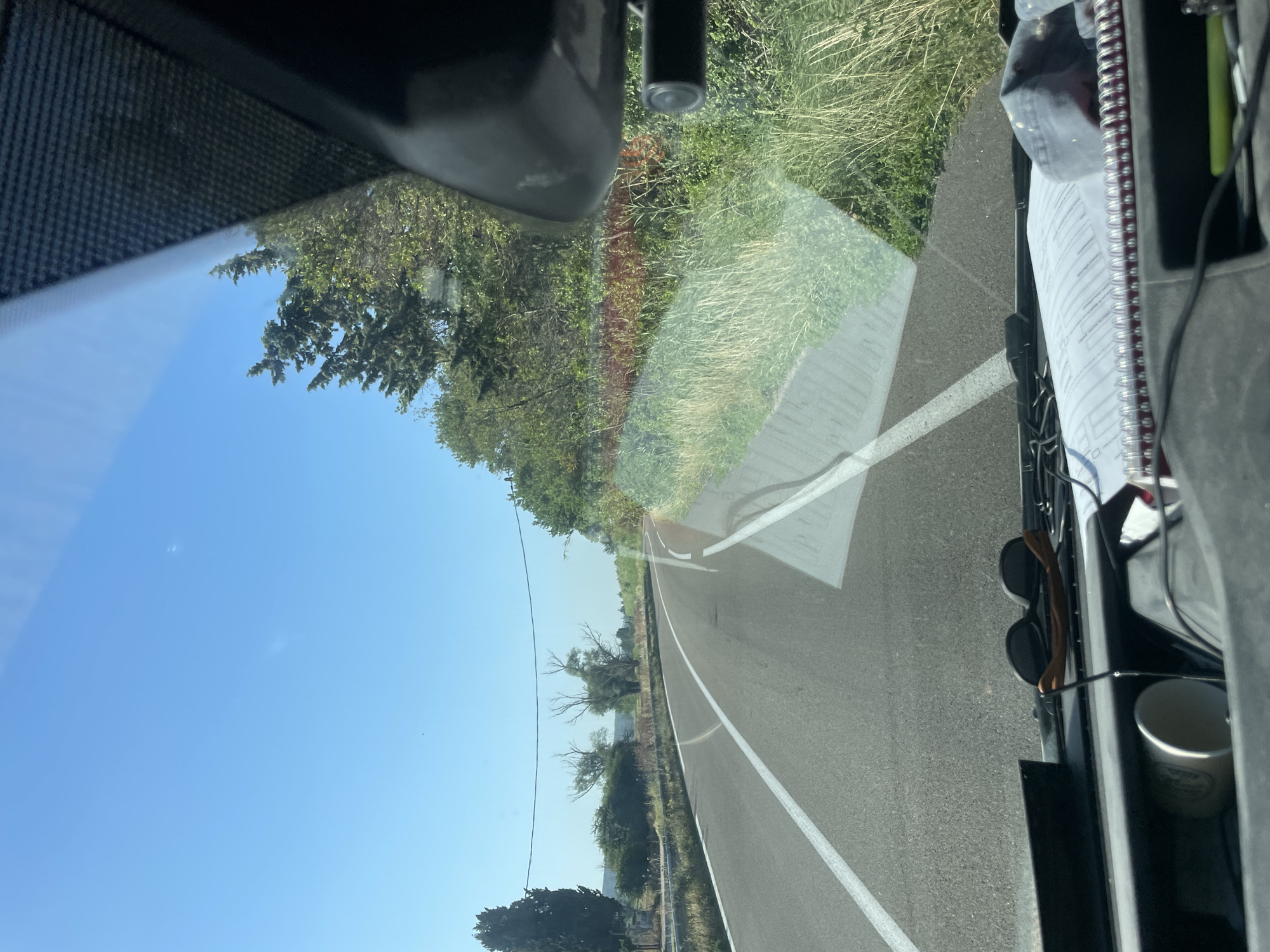 The image depicts a view from inside a vehicle looking forward through the windshield on a clear, sunny day. A two-lane asphalt road curves to the left, bordered by a white dashed line and a solid white line. On the right side of the road, tall grass and vegetation line the shoulder, with trees and foliage visible in the background. On the left side of the road, a line of trees and bushes can be seen, along with a single utility pole and a wire stretching across the sky. Reflections are visible on the windshield, showing parts of the vehicle's interior, including a rearview mirror with a small black camera attached, sunglasses resting on the dashboard, a white cup, and a notepad. The scene appears to be on a rural or suburban road, possibly on the outskirts of Tarascon, France, given the clear blue sky and Mediterranean-style vegetation.