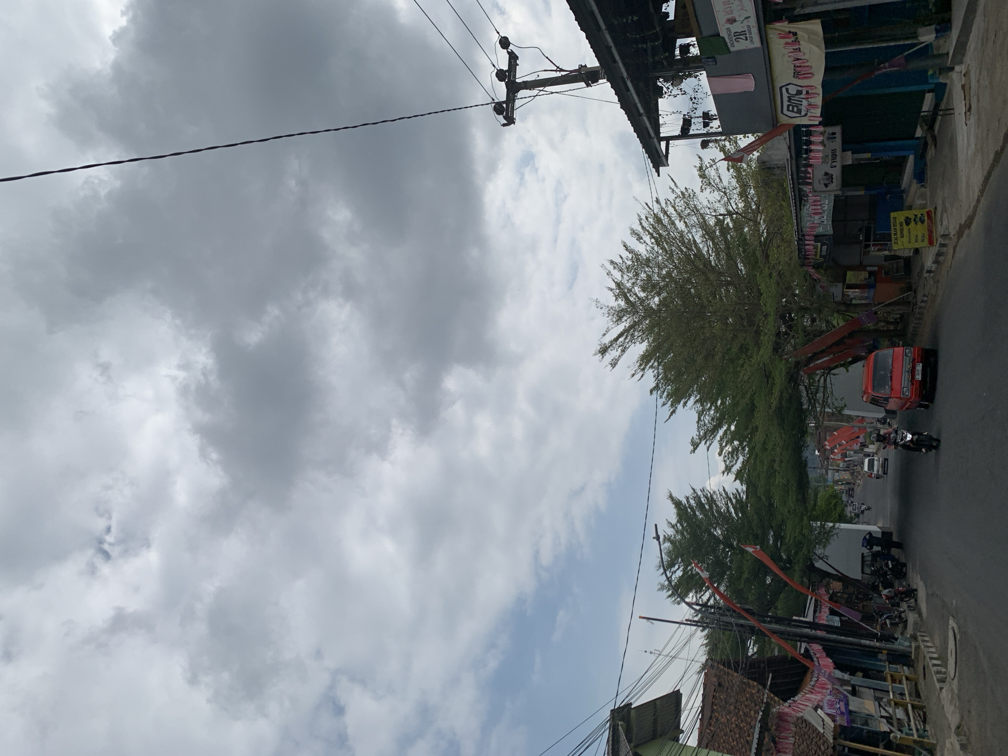 The image depicts a street scene in Ciamis, Indonesia, under a cloudy sky. The road is paved and appears to be in an urban or semi-urban area. On the left side of the street, there are buildings with tiled roofs and storefronts decorated with red and white banners, possibly for a celebration or national holiday. Several motorcycles are parked along the roadside. Further down the street, a red van is visible, and a person is riding a motorcycle. On the right side of the street, there are more buildings, including a structure with an overhang and hanging plants. Electrical poles and wires are prominent in the image, with one pole featuring multiple insulators. The trees lining the street have dense green foliage. The overall impression is a lively street with local commerce and transportation.