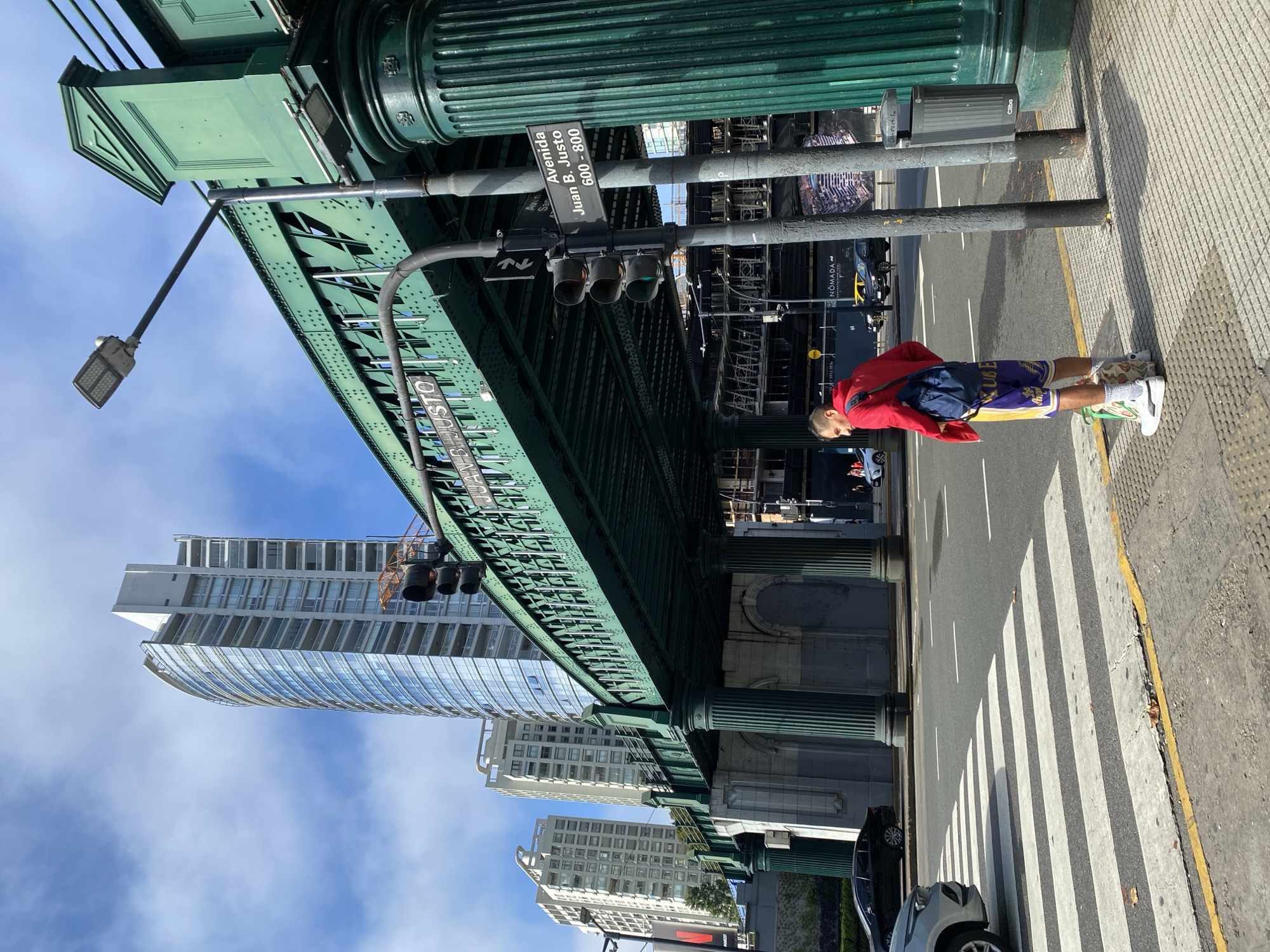 The image captures an urban scene in Buenos Aires, Argentina. An elevated green metal bridge, identified as part of the railway line over Avenida Juan B. Justo (between the 600 and 800 block), dominates the foreground. The street runs parallel to the bottom edge of the image, with a marked pedestrian crossing and parked cars along the curb. A man in a red jacket and purple and gold shorts stands on the sidewalk near a metal trash can. Tall buildings rise in the background, partially obscured by the bridge structure. The sky is blue with scattered clouds. Traffic lights hang from the bridge structure, positioned above the street.
