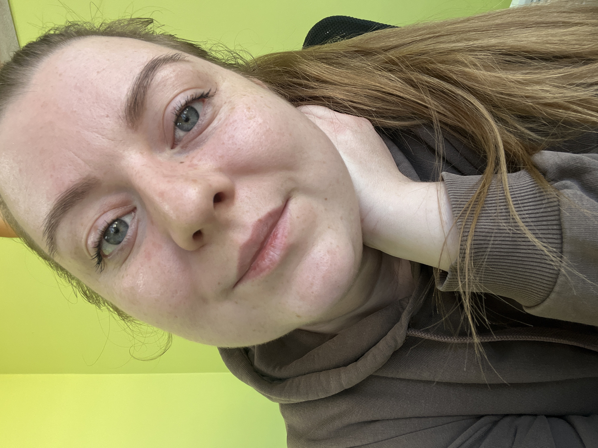 A close-up portrait features a female individual, appearing adult, situated in an interior location in Klin, Russia. She possesses light skin with visible freckles across her face, blue eyes, and light brown hair, which is pulled back with strands visible on the right side of the frame. Her left hand is positioned supporting her chin and lower jaw. She is wearing a dark olive-green or brown hooded sweatshirt with ribbed cuffs. The subject is looking directly towards the camera, displaying a subtle smile. The background is a solid, bright lime-green wall, with an interior corner visible towards the left. A dark, indistinct object is partially visible behind her right shoulder.