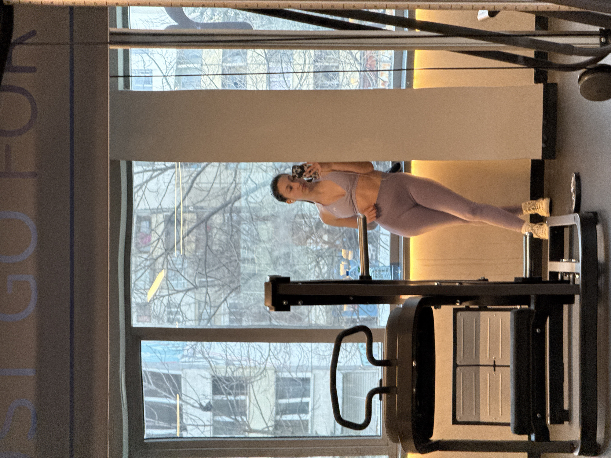 The scene is indoors, likely a gym, in front of a large window with a view of a cityscape. The buildings outside suggest an urban environment, potentially in Sofia, Bulgaria, although this would require local knowledge for confirmation. A woman in athletic wear (light purple sports bra and leggings) is visible, holding a smartphone in her hand as if taking a selfie in front of a workout machine. The machine appears to be a leg press or similar exercise equipment. The overall lighting is bright, with natural light coming through the window. The gym wall to the left has text, potentially motivational, partially visible.