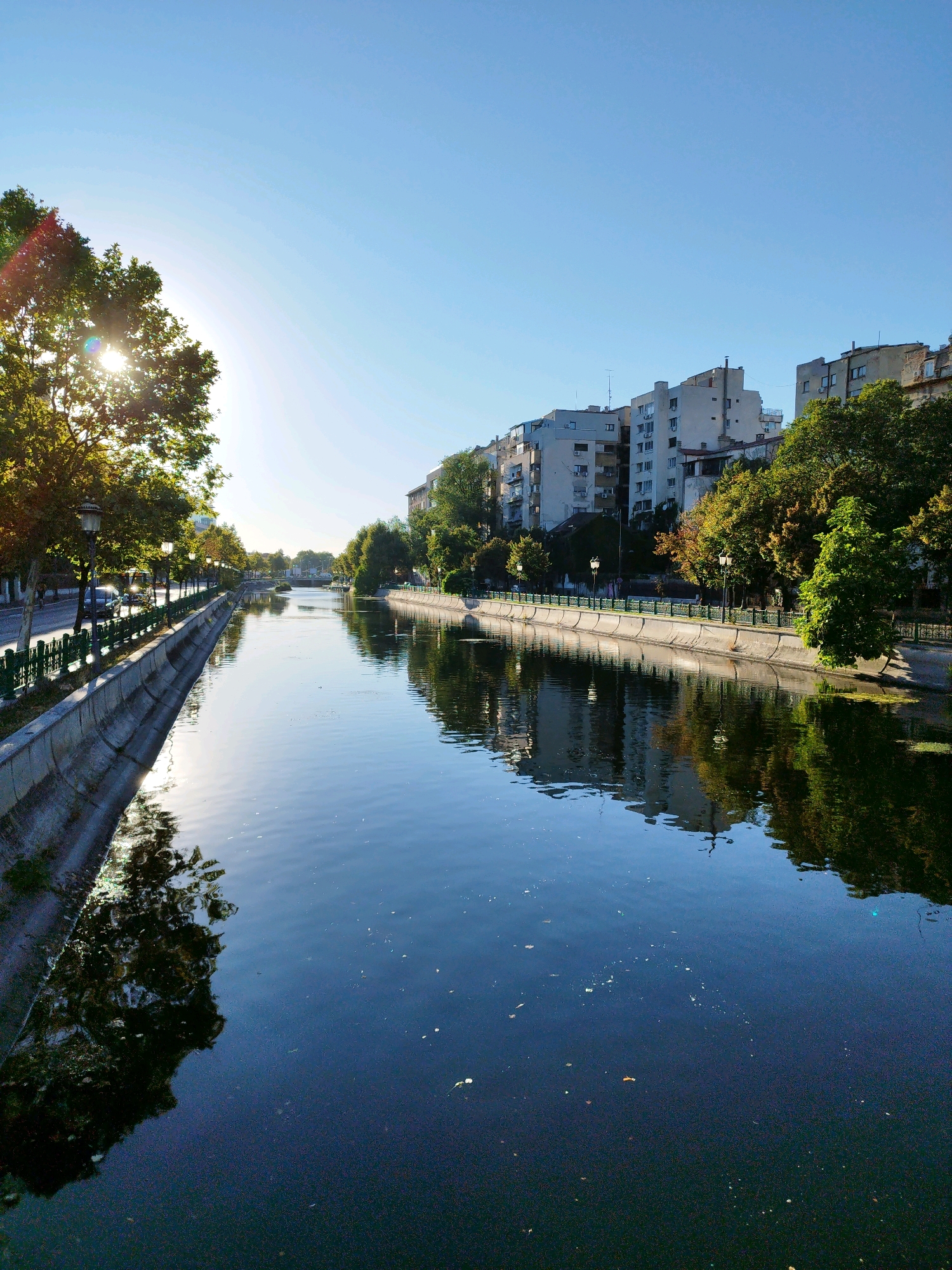 This image captures a view of the Dâmbovița River in Bucharest, Romania, likely in the morning given the low angle of the sun and the clear, bright sky. The river flows through an urban landscape, with concrete embankments lining its sides. On the left bank, a tree-lined street is visible, with lampposts and cars suggesting a typical city thoroughfare. The buildings across the river on the right bank are multi-story residential structures, some appearing to be older Soviet-era apartment blocks. The water reflects the sky and the surrounding environment, with some debris visible on the surface. The sun is shining brightly, creating a lens flare effect in the upper left portion of the image and casting reflections on the water and trees. The overall scene depicts a tranquil urban waterway on a sunny day.