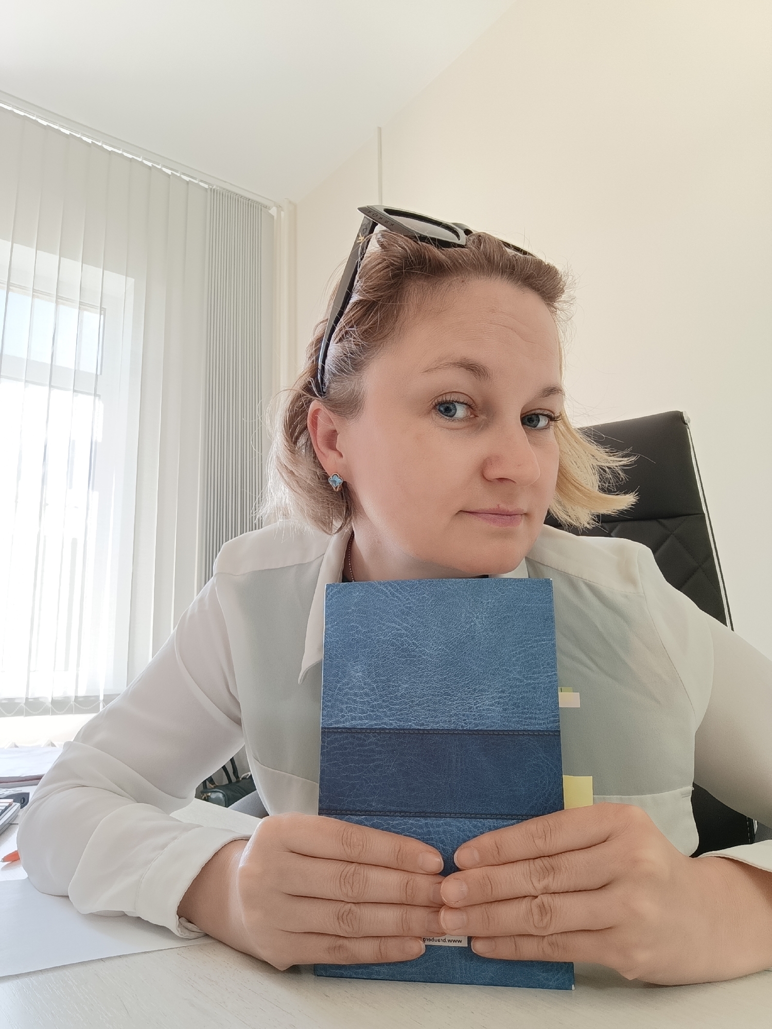 A medium shot shows a woman with blonde hair wearing a white shirt sitting at a desk in an office. She has sunglasses perched on her head and is holding a blue notebook with both hands in front of her. Her right arm is resting on the desk, and the notebook is positioned in front of her chest. The office has white vertical blinds on a window to the left and a black office chair behind the woman. The desk surface is visible in the foreground. The scene appears to be in the city of Марковский, Russia.