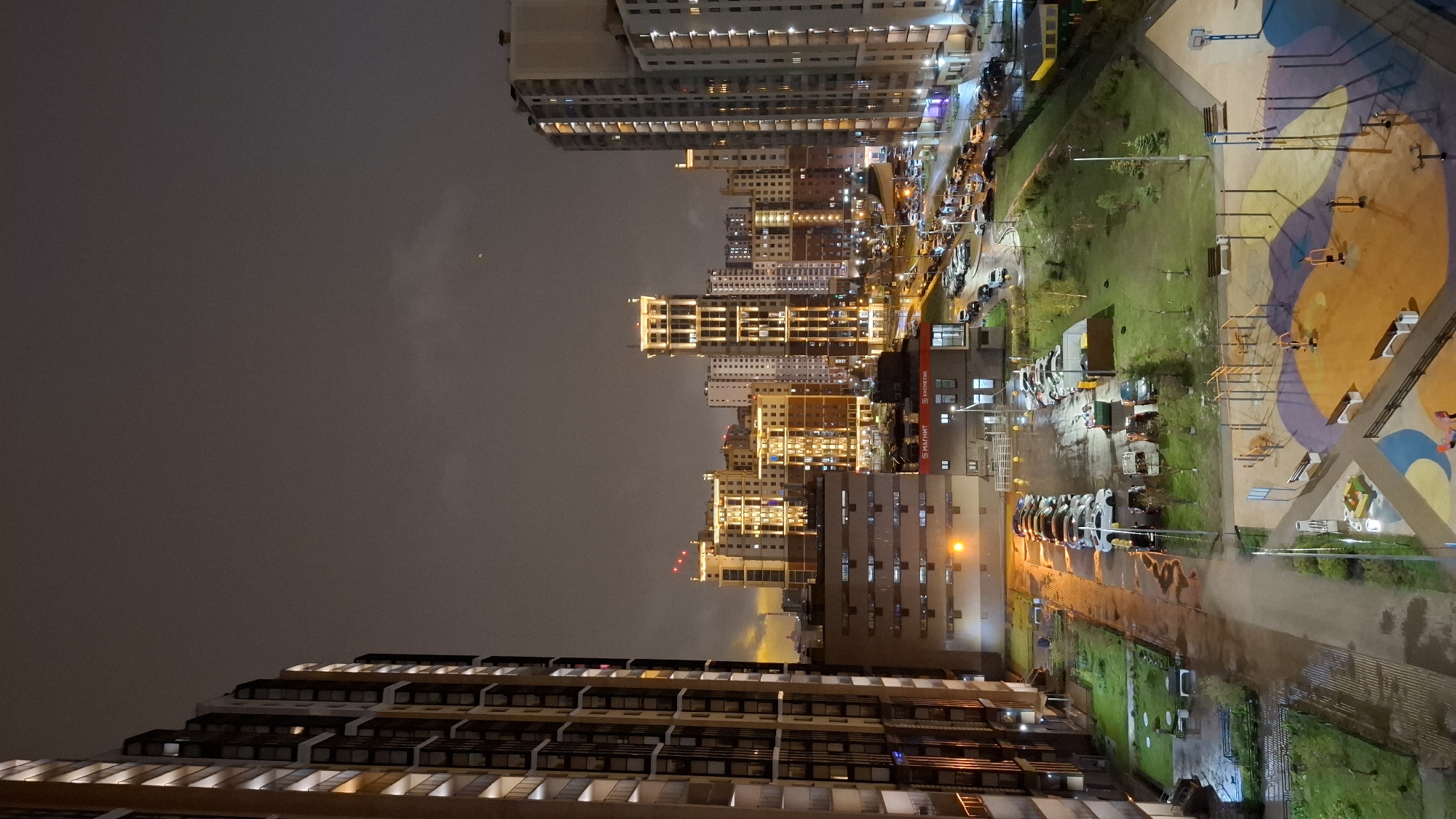 An aerial view of Saint Petersburg, Russia, taken at night shows a city skyline and a playground. The city buildings are lit with warm yellow lights, contrasting with the dark grey sky. The playground features colorful surfaces, grass, and exercise equipment. Cars are parked along a street adjacent to the playground.