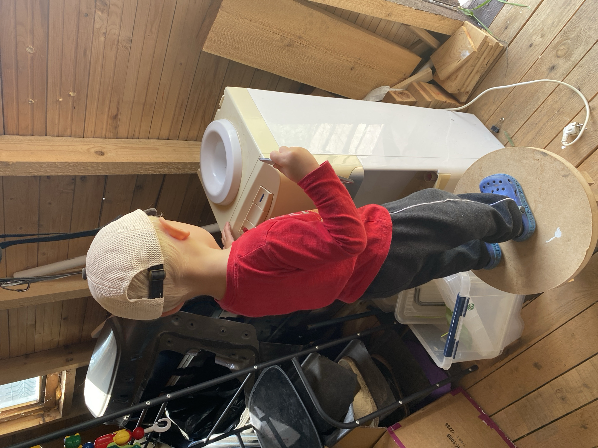 A young child wearing a red long-sleeved shirt, black pants with a white stripe, blue Crocs, and a light-colored mesh baseball cap stands on a round wooden stool. The child is facing away from the camera, towards a white and beige water cooler. The child is holding a silver object, possibly a utensil, near the top of the water cooler. The water cooler has a white dispenser area at the top. The child is positioned in a rustic wooden room with wooden plank walls and floor. To the left of the water cooler is a dark metal object with black components, resembling part of a disassembled appliance or equipment. In the background, a window with sunlight streaming through is visible. There are various wooden planks and objects stacked on the right side of the image. The overall environment appears to be a storage area or workshop with a casual, slightly cluttered feel.