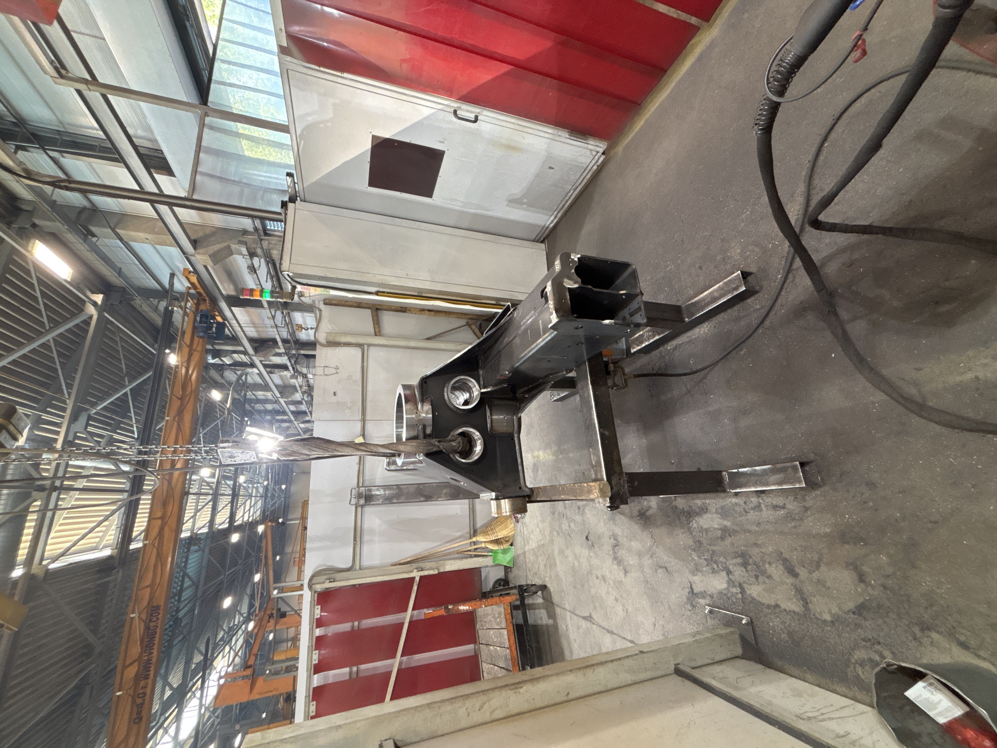In the industrial city of Ruptsi, Bulgaria, within a large manufacturing facility, a heavy metal component is suspended by a crane. The component, which appears to be part of a machine or vehicle, is dark gray and features several circular openings. It is held by a metal cable and hook assembly. The environment is a concrete floor with various industrial elements visible, including support beams, lighting fixtures, red industrial curtains, a trolley with equipment, and brooms. Electrical cables snake across the floor, suggesting ongoing work. The scene is brightly lit, likely by overhead industrial lighting and natural light filtering through large windows or skylights. The overall impression is one of active industrial production.