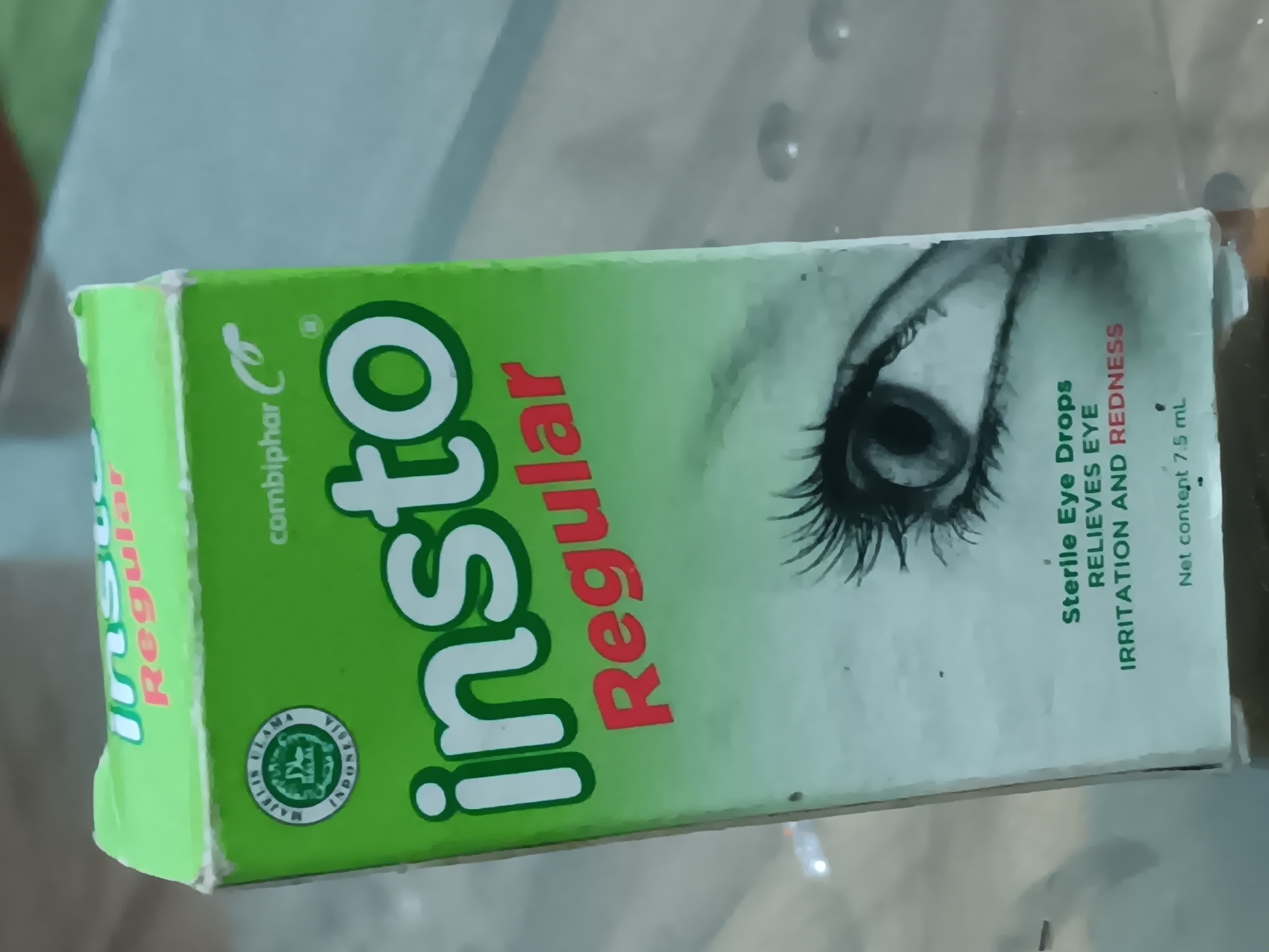 The image shows a close-up of a green cardboard box for "Insto Regular" eye drops. The box features the brand name "Insto" in large white letters with a green outline, and "Regular" in red letters below it. A stylized drawing of an eye with eyelashes is prominently displayed on the lower half of the box. Text on the box indicates "Sterile Eye Drops," "RELIEVES EYE IRRITATION AND REDNESS," and "Net content 7.5 mL." A circular halal certification logo is visible on the upper left side of the box. The box appears slightly worn, with some creasing and minor damage to the edges, suggesting it has been handled or stored for some time. The background is out of focus, but suggests an indoor setting, possibly a table surface, with reflections and lighting that hint at a domestic or retail environment. The context of Batursari, Indonesia, is inferred from the "Majelis Ulama Indonesia" text on the halal logo. There are no people or actions depicted in the image.