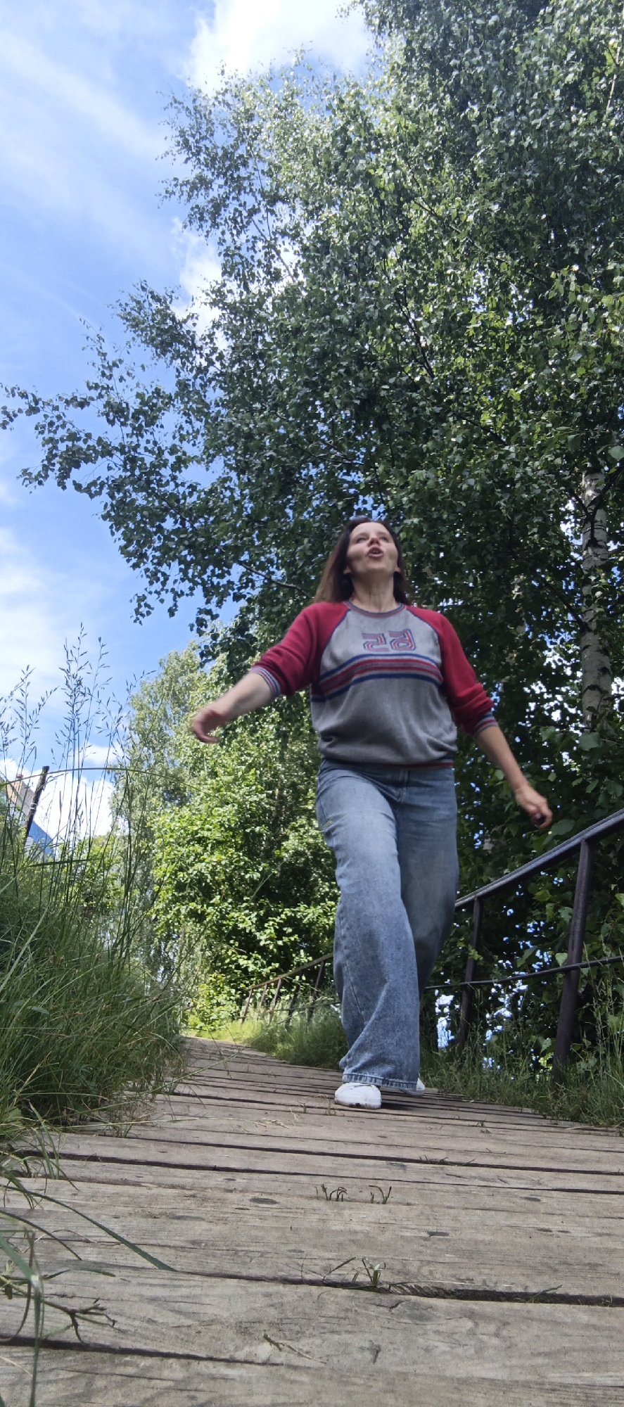 In an outdoor, low-angle shot, a young woman with medium-length brown hair walks down a wooden boardwalk surrounded by lush green trees and a bright blue sky with scattered clouds. She is wearing a grey long-sleeved shirt with red and blue stripes on the chest, and loose-fitting blue jeans. Her arms are outstretched as she walks, and her mouth is open in a singing or yelling expression. The boardwalk appears weathered, with visible planks and grass growing along the sides. A dark metal railing is present on the right side of the boardwalk. The overall scene depicts a moment of playful exuberance in a natural, possibly park-like, setting.