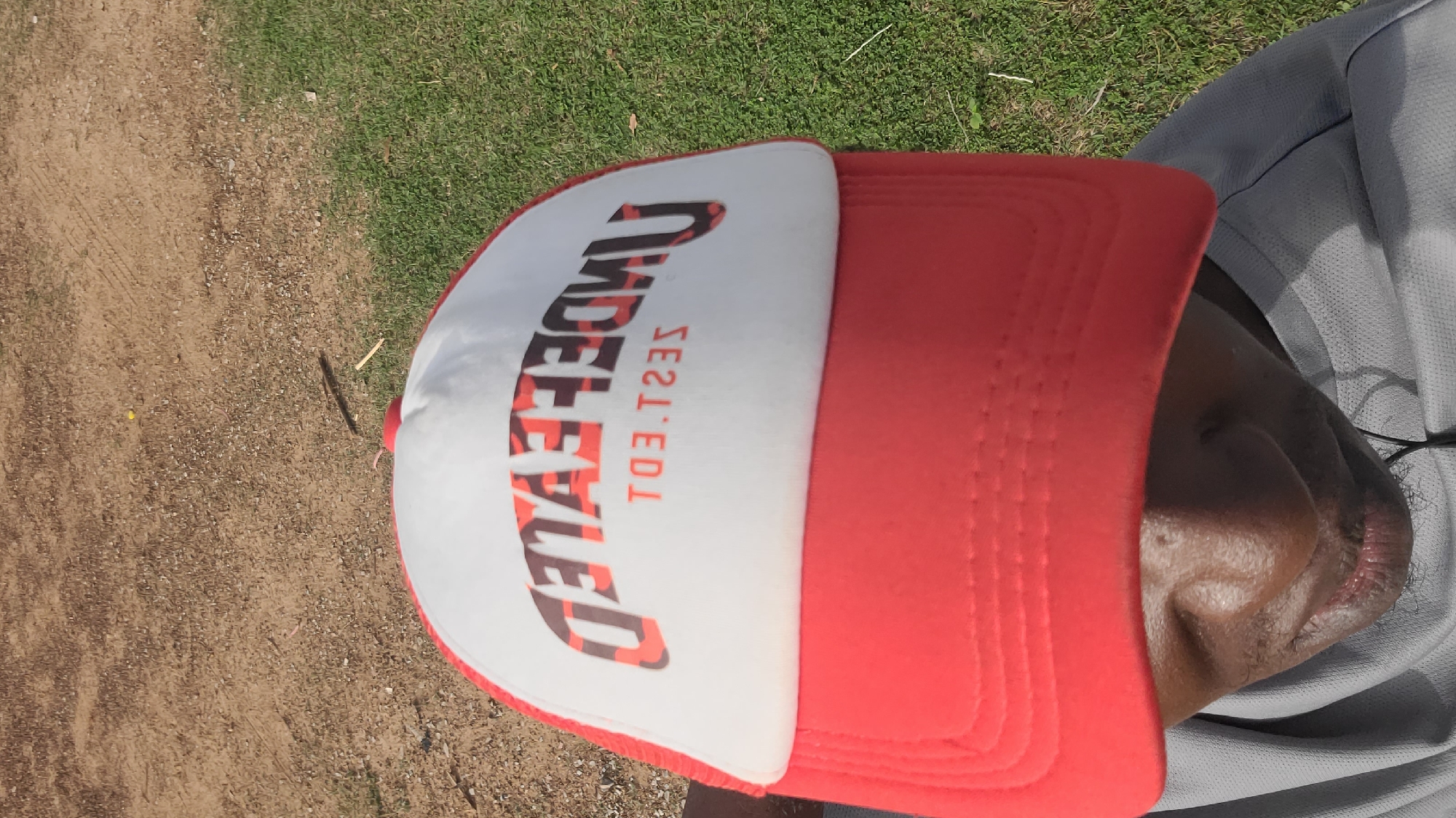 A person wearing a red and white baseball cap with the word "UNDEFEATED" printed on it is positioned in a grassy area adjacent to a dirt path. The person is wearing a gray shirt. The location appears to be outdoors in daylight. Specific details to identify the location as the city of Ogbomosho, Nigeria are missing from the image.