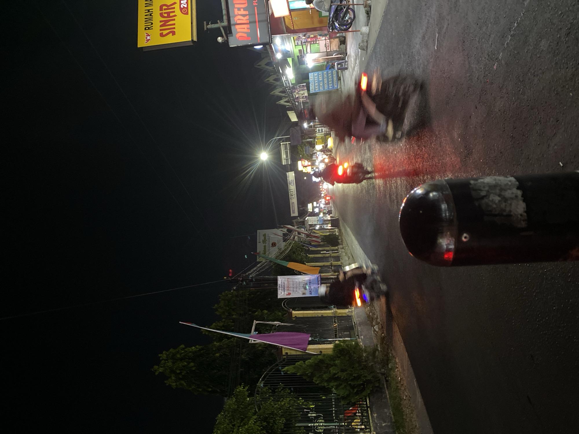 The image depicts a nighttime street scene in Kasihan, Indonesia, characterized by low light conditions and motion blur from moving vehicles. A street lined with businesses is visible, with glowing signs indicating various establishments. One sign, yellow with red lettering, reads "RUMAH MA SINAR" and includes a phone number with a "24" prefix, suggesting a 24-hour service. Another sign, with red lettering on a dark background, says "PARFU" which is likely short for perfume shop. In the foreground, a dark cylindrical object, possibly a bollard or a post, is partially visible. The road surface appears wet, reflecting the lights. Several motorcycles are in motion, indicated by blurred shapes and illuminated taillights. One motorcycle in the right midground is significantly blurred, with its rider wearing a light-colored garment. Another motorcycle in the left midground is also blurred, with its red taillight clearly visible. Further down the street, more headlights and taillights indicate additional traffic. On the left side of the street, a black metal fence borders a property with trees and flags, one of which is purple and white. A banner with text, including "ARCHITECT," is attached to the fence. Utility poles with numerous wires are present along the street. The overall impression is a busy urban street at night.
