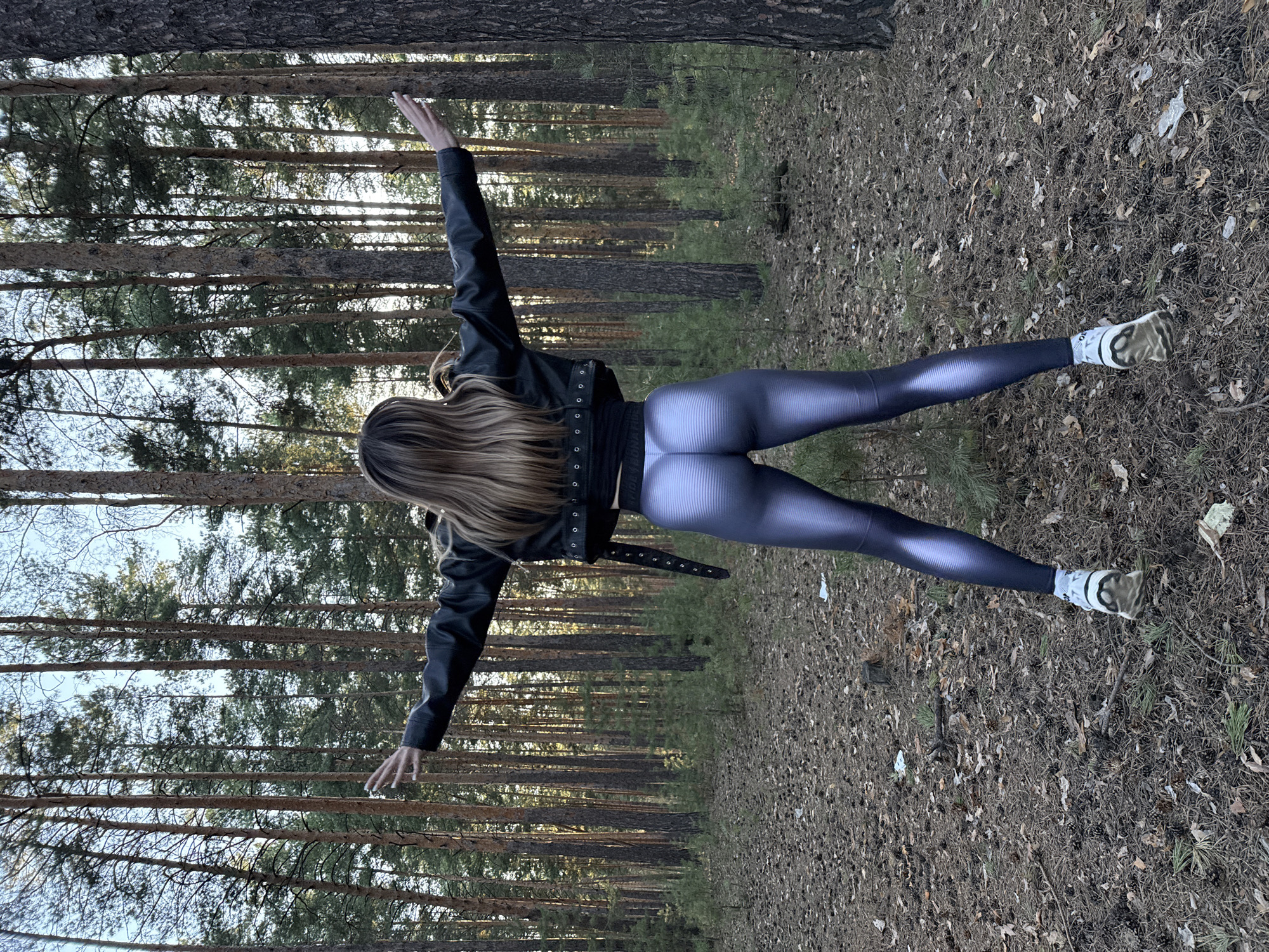An overhead, rotated full shot captures a person from behind, standing in a pine forest. The individual has long, light brown hair and is dressed in a black leather jacket, a dark top, and reflective dark grey/purple athletic leggings with a subtle horizontal ribbed pattern. White athletic socks, stained with dirt and featuring a dark logo, are visible, along with white and beige sneakers that also appear soiled.
The person strikes a balanced pose, with arms extended horizontally to the sides and one leg lifted backward, suggesting a moment of exercise, stretch, or expressive movement within nature. The scene is set in a dense forest, characterized by numerous tall, slender pine trees with textured, reddish-brown bark that stretch upwards. Sunlight filters through the tree canopy, creating a dappled effect of light and shadow on the scene.
The forest floor is covered with a natural layer of fallen pine needles, small branches, and dry leaves, with occasional patches of sparse green undergrowth. The overall impression is one of an outdoor activity in a serene woodland environment, likely during daylight hours, possibly late afternoon or early morning, given the quality of light. No specific text is clearly legible on any item of clothing beyond a generic logo on the socks.