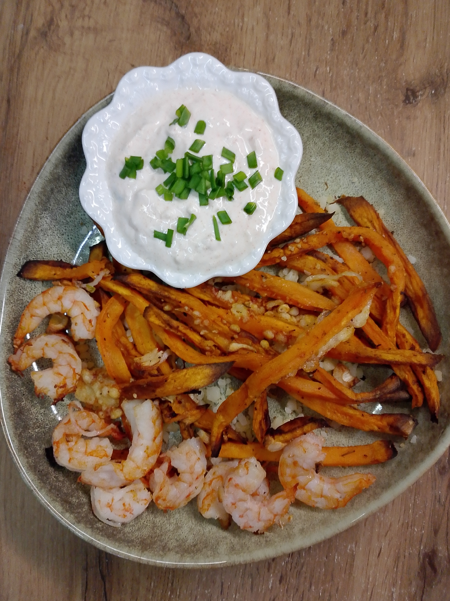 A top-down shot shows a plate of food on a wooden surface. The plate contains sweet potato fries sprinkled with a granular substance, cooked shrimp, and a small white bowl filled with a creamy sauce garnished with chopped chives. No information suggests this is in Izhevsk, Russia.