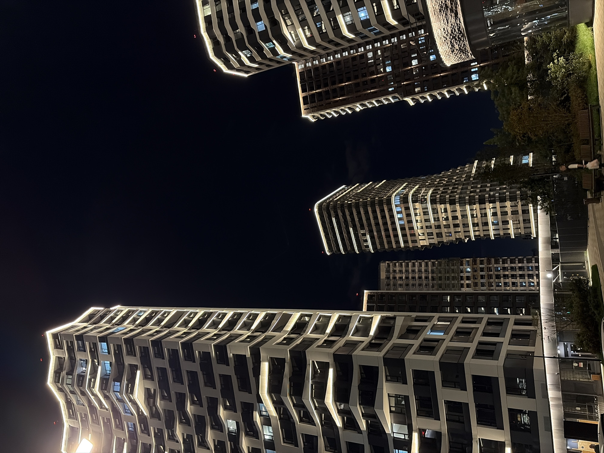 The image depicts a nighttime view of the residential complex "Spires of Metropolis" in Moscow, Russia. Several modern high-rise buildings with illuminated facades are prominently featured against a dark night sky. The architecture is characterized by angular designs and extensive use of glass and light accents along the edges of the structures. In the foreground, a walkway with benches and some greenery is visible. A person walking with a bag and a dog is present on the walkway, and another person is seated on a bench. The lighting from the buildings creates a dramatic and urban atmosphere.