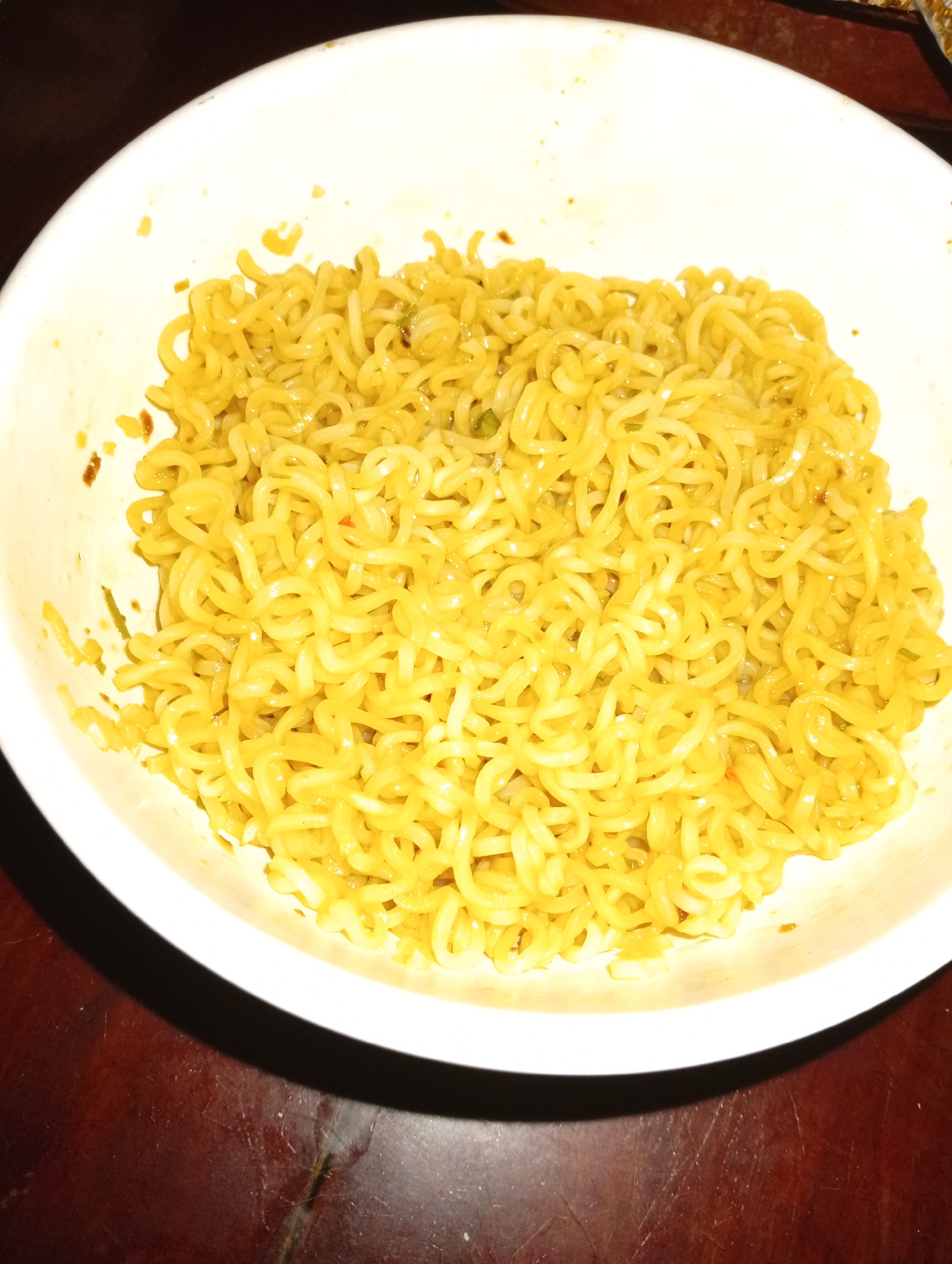 A close-up, overhead shot shows a bowl of yellow instant noodles. The noodles are coiled and appear to be seasoned with spices. The white bowl is slightly chipped on the left side. The noodles fill most of the bowl, which is placed on a dark wooden surface. The lighting is dim, casting a shadow on the lower left side of the bowl. The location is within the city of Cordon, Philippines.