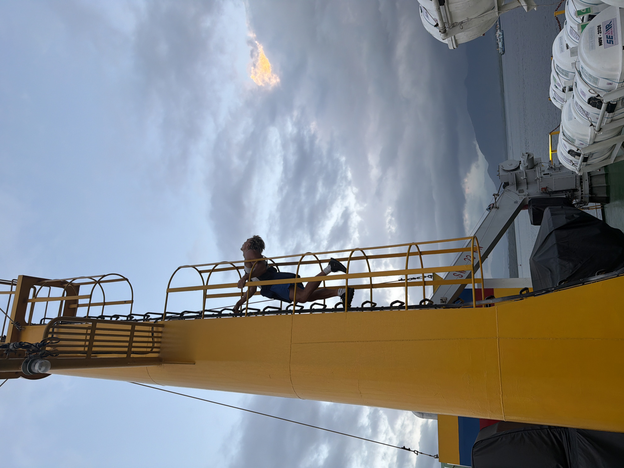 The image shows a person climbing a yellow ladder on the side of a ship. The person, dressed in shorts and a t-shirt, is reaching up to grab the next rung. The ship appears to be docked or at sea, with a body of water and mountains visible in the background. The sky is cloudy with some areas of sunlight breaking through. Lifeboats are visible on the deck of the ship. The location is identified as Melaya, Indonesia, suggesting the ship may be in a port or harbor in that region.