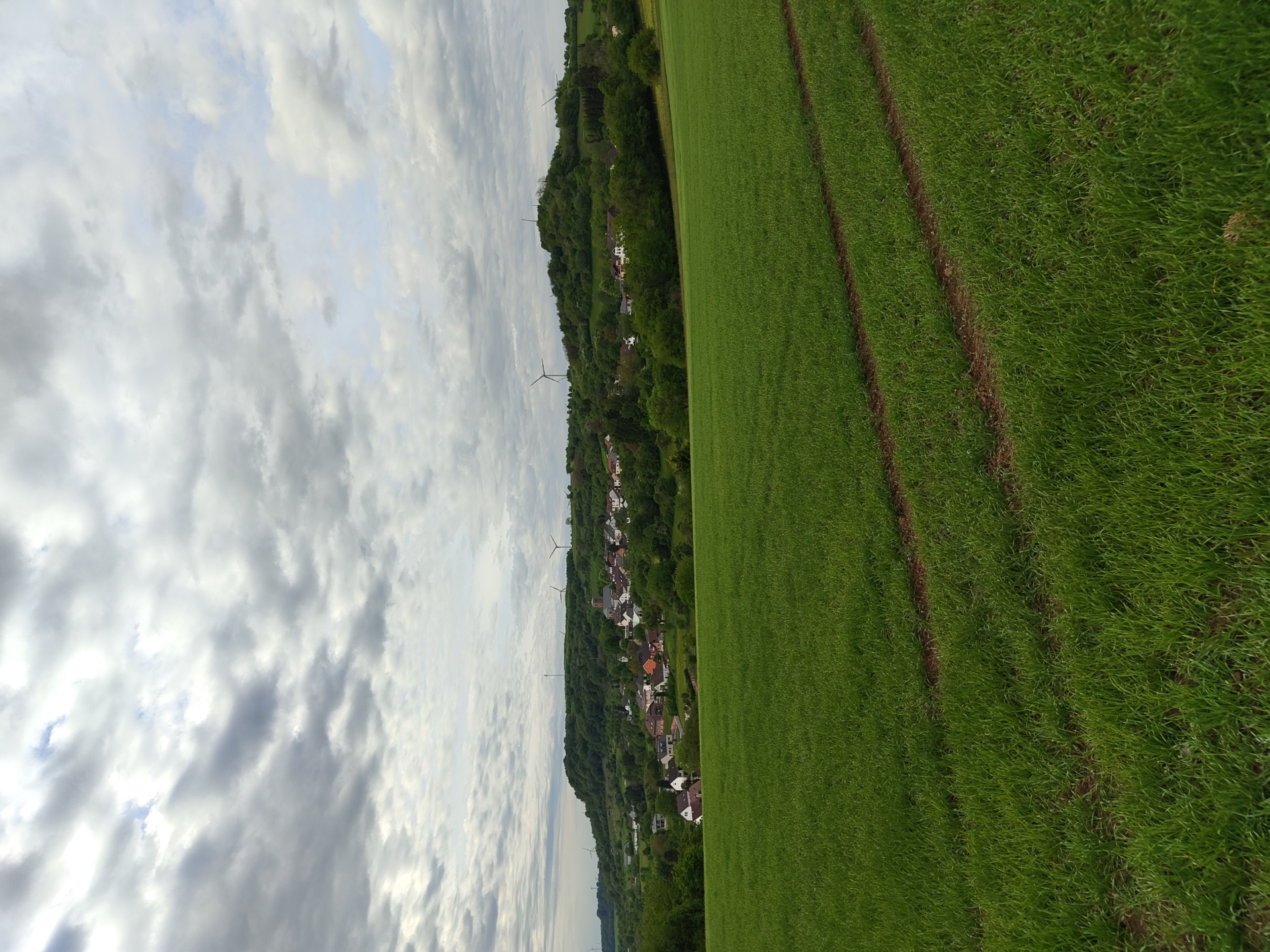 The image shows a landscape in Perl, Germany. A large field of green grass occupies the right side of the frame, with dark brown tracks running through it. To the left of the field, a tree-covered hill leads down to a village with houses that have red and white roofs. Several wind turbines are visible along the ridge of the hill. The sky above is filled with numerous gray and white clouds.