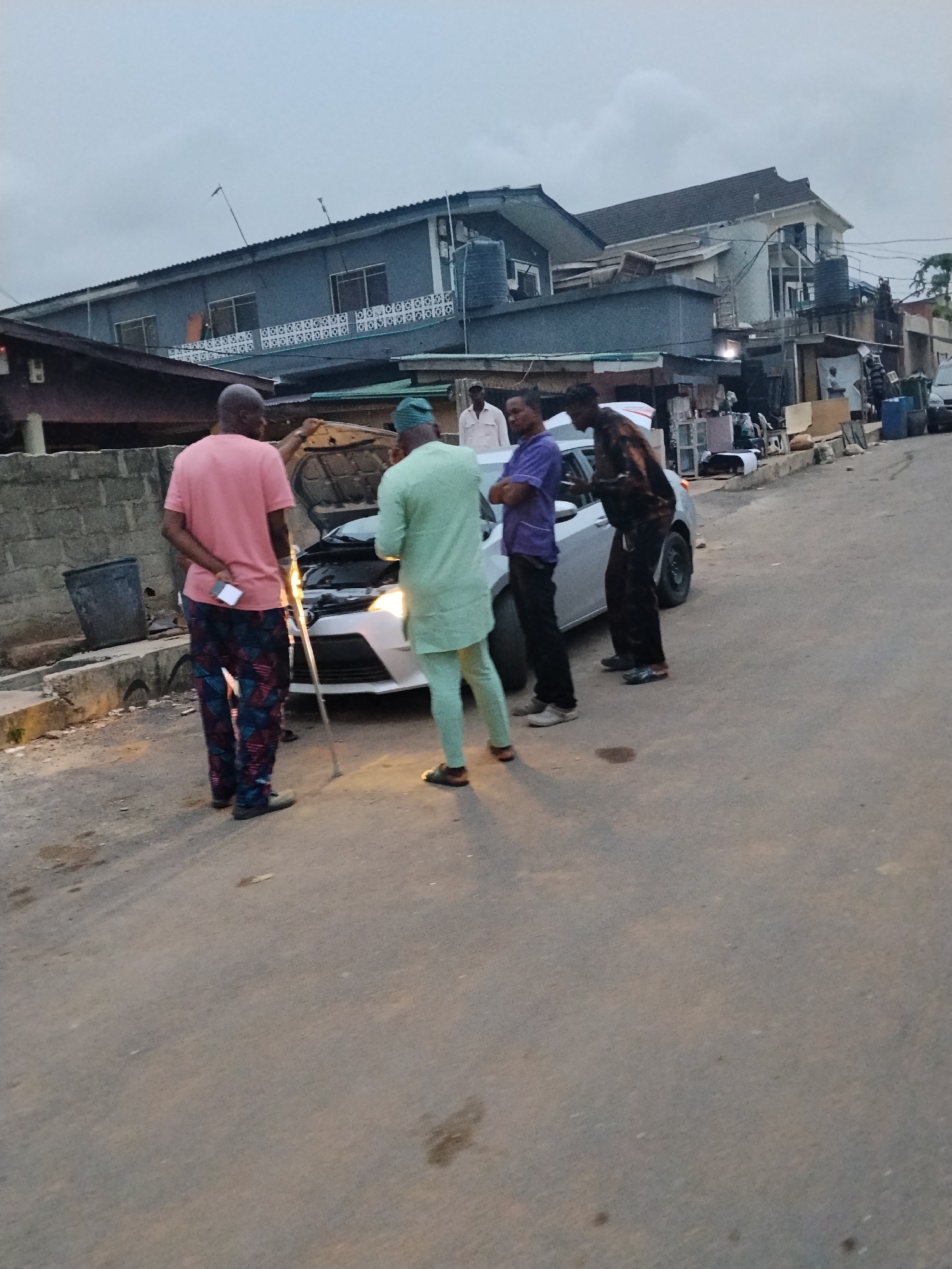 This image depicts a roadside scene in Shangisha, Nigeria, where a group of men are gathered around a white car with its hood open, suggesting a breakdown or inspection. The car's headlights are illuminated, indicating it is likely late afternoon or early evening under an overcast sky.
Five men are visible. In the foreground, a man in a pink t-shirt and patterned trousers stands with his back to the viewer, leaning on a crutch. Next to him, a man in a light green traditional kaftan is looking into the car's open hood. To their right, a man in a purple t-shirt stands with his arms crossed, observing the car. Another man in a dark, patterned top and dark trousers also stands near the car, looking towards the engine. A fifth man, wearing a light-colored shirt, is partially visible further back, behind the man in green.
The setting is an urban street, characterized by an asphalt road with visible stains. Along the left side of the road, there is a low concrete wall with a black bin. In the background, multiple buildings line the street, featuring corrugated iron roofs, some with water tanks on top. The structures appear to be a mix of residential and ground-level commercial establishments, with various items like furniture pieces and blue barrels scattered along the roadside. The overall mood is focused and uneventful, centered on the car's issue. No visible text can be clearly identified.