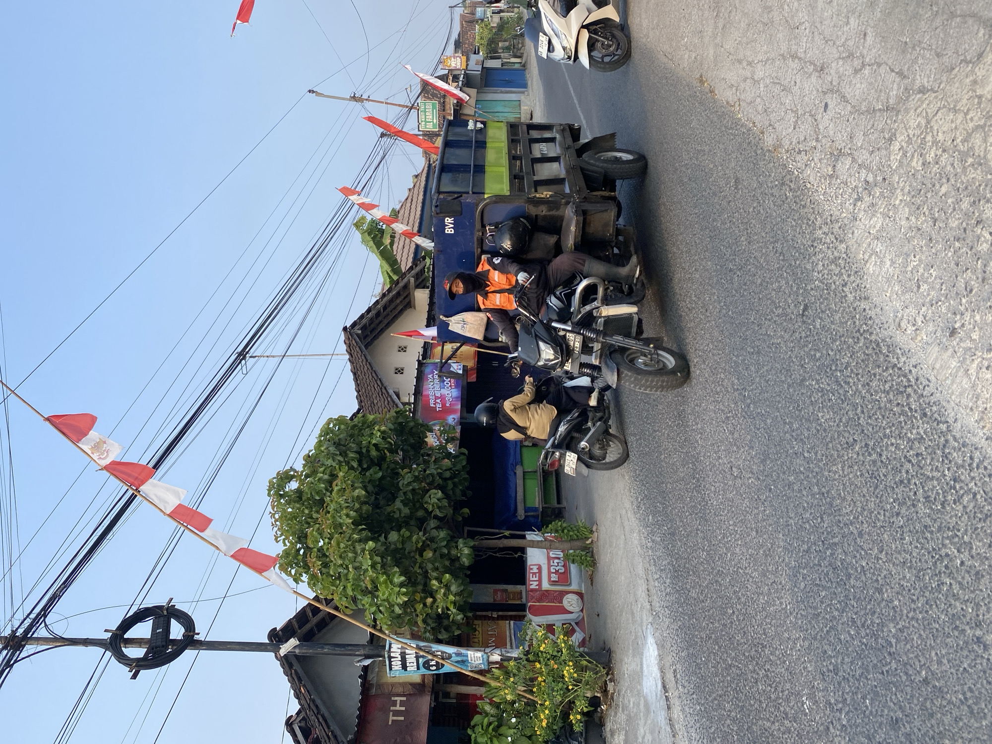 The image captures a street scene in Kasihan, Indonesia, decorated with Indonesian flags, possibly in celebration of Independence Day. In the foreground, a three-wheeled vehicle, a modified motorcycle with a cargo bed, is driven by a person wearing a face mask and an orange vest. The cargo bed is filled with dark blue containers. To the left of the three-wheeled vehicle, a person on a motorcycle is also visible. In the background, buildings line the street, and overhead, a complex network of electrical wires is strung between poles. The road surface is asphalt, and there are scattered plants and trees along the roadside. The sky is clear and blue.