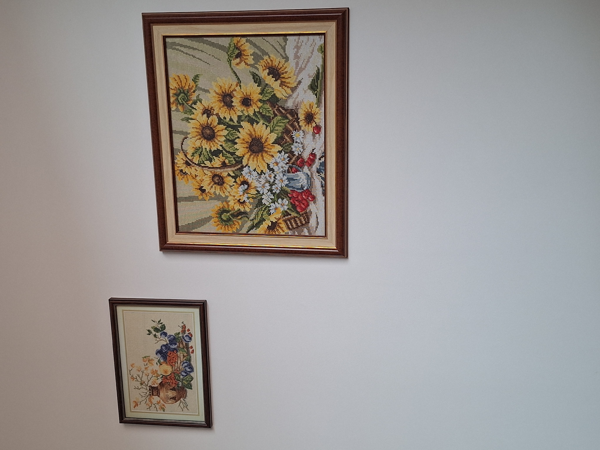 The image shows two framed cross-stitch pictures hanging on a white wall. The larger picture depicts a bouquet of sunflowers and other flowers in a basket. The smaller picture shows a vase of blue and orange flowers. The location is likely indoors, but there is no information about the specific location in Montana, Bulgaria. There are no people in the image.