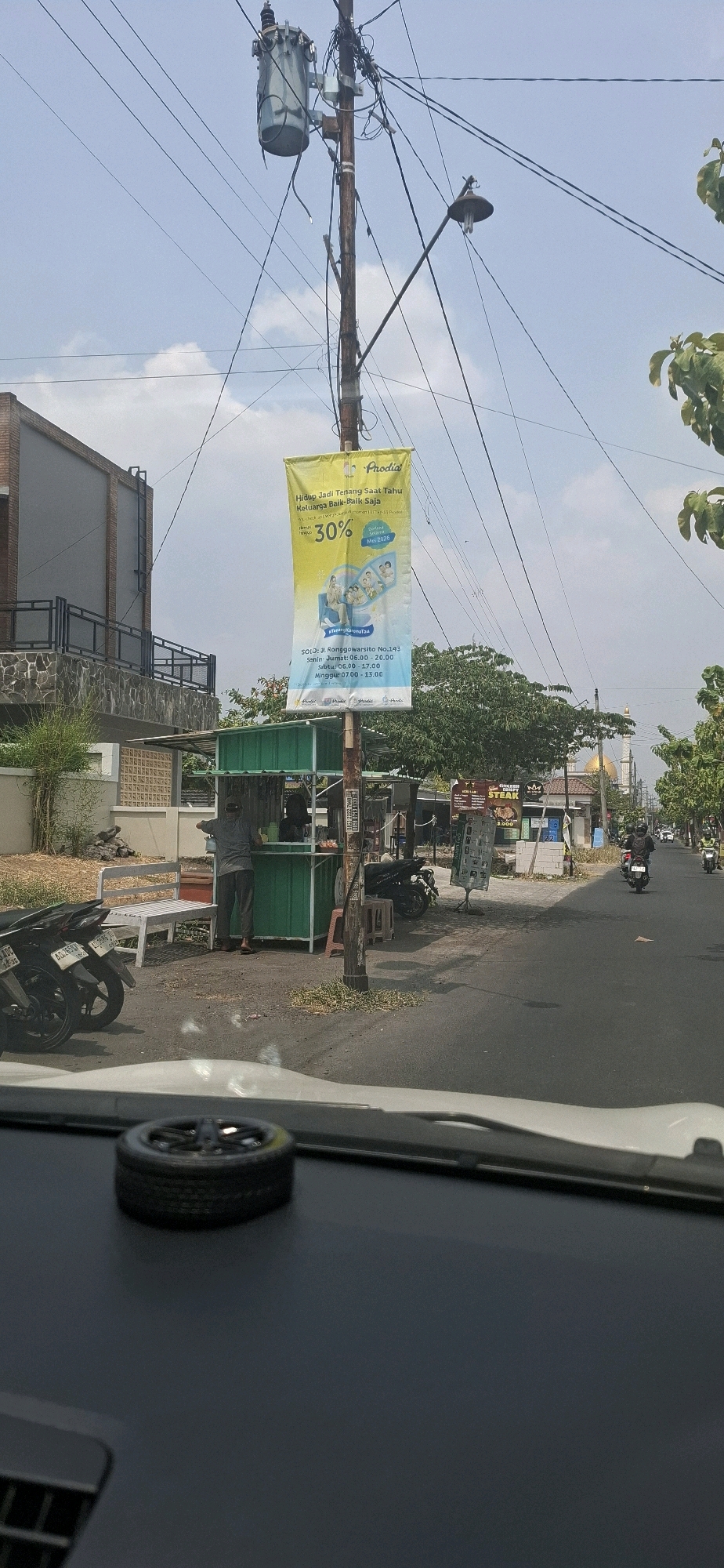 A bright yellow banner hangs from a utility pole near a food stall in Pabelan, Indonesia. The banner advertises a promotion with "30%" off from Prodia, a healthcare company, and mentions "Hidup Jadi Tenang Saat Tahu Keluarga Baik-Baik Saja." The food stall is green and has a white bench in front of it. A person in a gray shirt and dark pants is standing at the stall. Several motorcycles are parked nearby. The street appears to be a local road, with buildings and trees lining one side. The sky is blue with some clouds. The foreground shows the dashboard of a car with a tire-shaped air freshener.