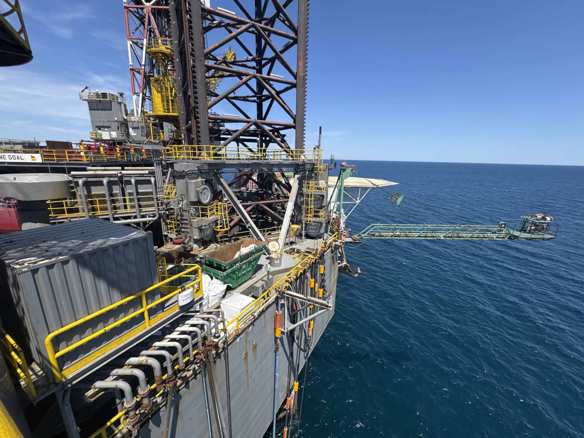 This media file depicts an offshore drilling rig on a clear, sunny day. The rig, a massive industrial structure, dominates the frame with its metal framework, pipes, and various platforms. Several large containers, including a gray corrugated metal one and a green dumpster, are visible on one of the decks. Yellow safety railings line the edges of the platforms.
The rig is situated on a vast expanse of deep blue ocean. The horizon is clearly defined where the water meets the bright blue sky, with no land in sight. A small red boat is visible in the distance on the water. A helideck extends from one side of the rig, suggesting it is accessible by helicopter.
The overall impression is one of industrial operation in a remote maritime environment. There is no visible text in the frame, except for what appears to be some labeling on the gray container, which is too small to be legible. The lighting suggests it is daytime, likely midday, given the bright sun and clear sky. The image conveys a sense of isolation and the immense scale of industrial offshore operations.