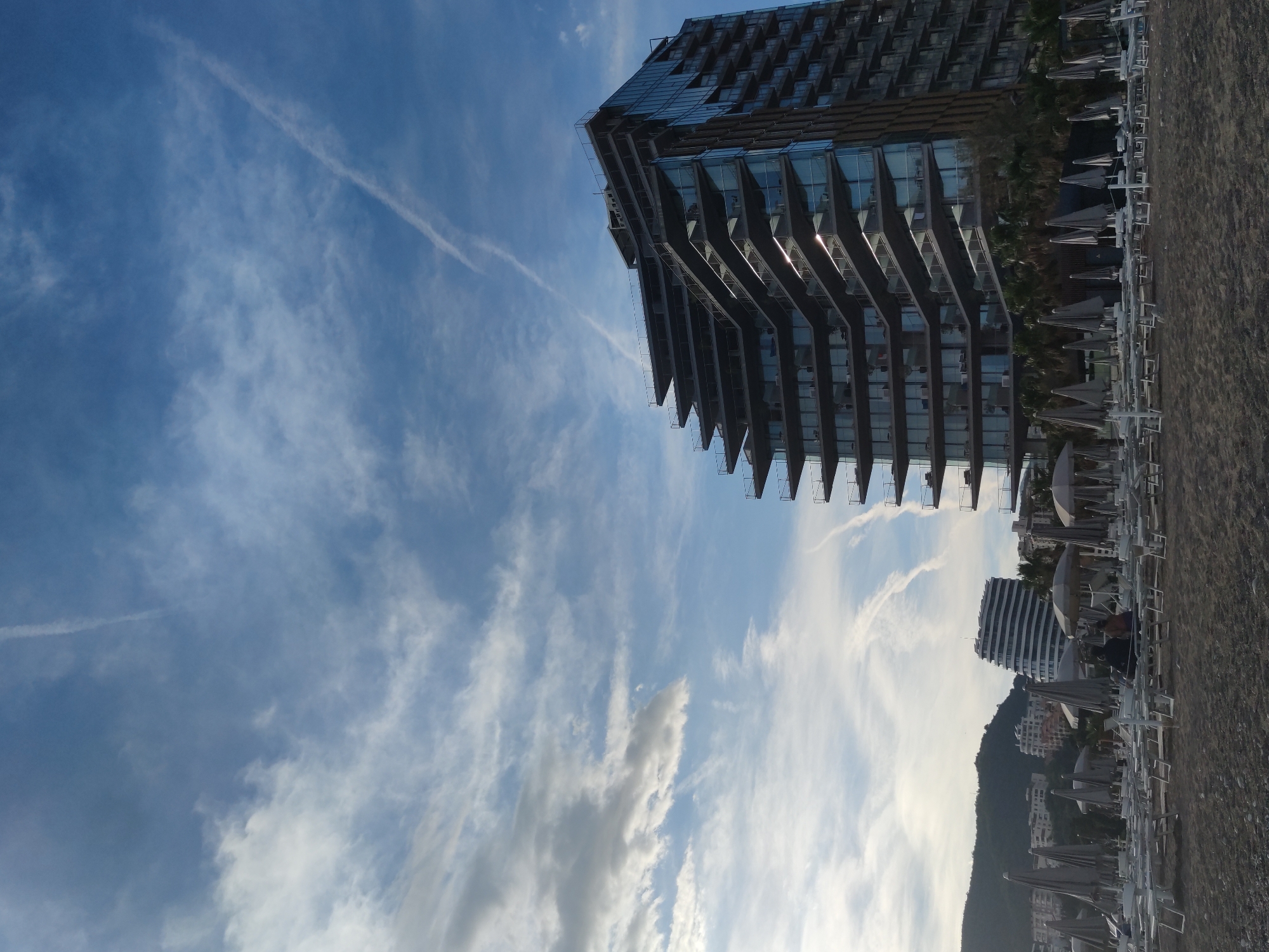 The image shows a beach scene with buildings. In the top right, a tall modern building has a distinctive angular design. To the right of it, there is a row of closed white beach umbrellas and chairs along the sandy beach. In the background, there's a smaller white building, a green mountain, and other buildings. The sky above is blue with wispy clouds and contrails from an airplane. The image appears to be rotated to the side. The location is in the city of Unknown, Unknown.